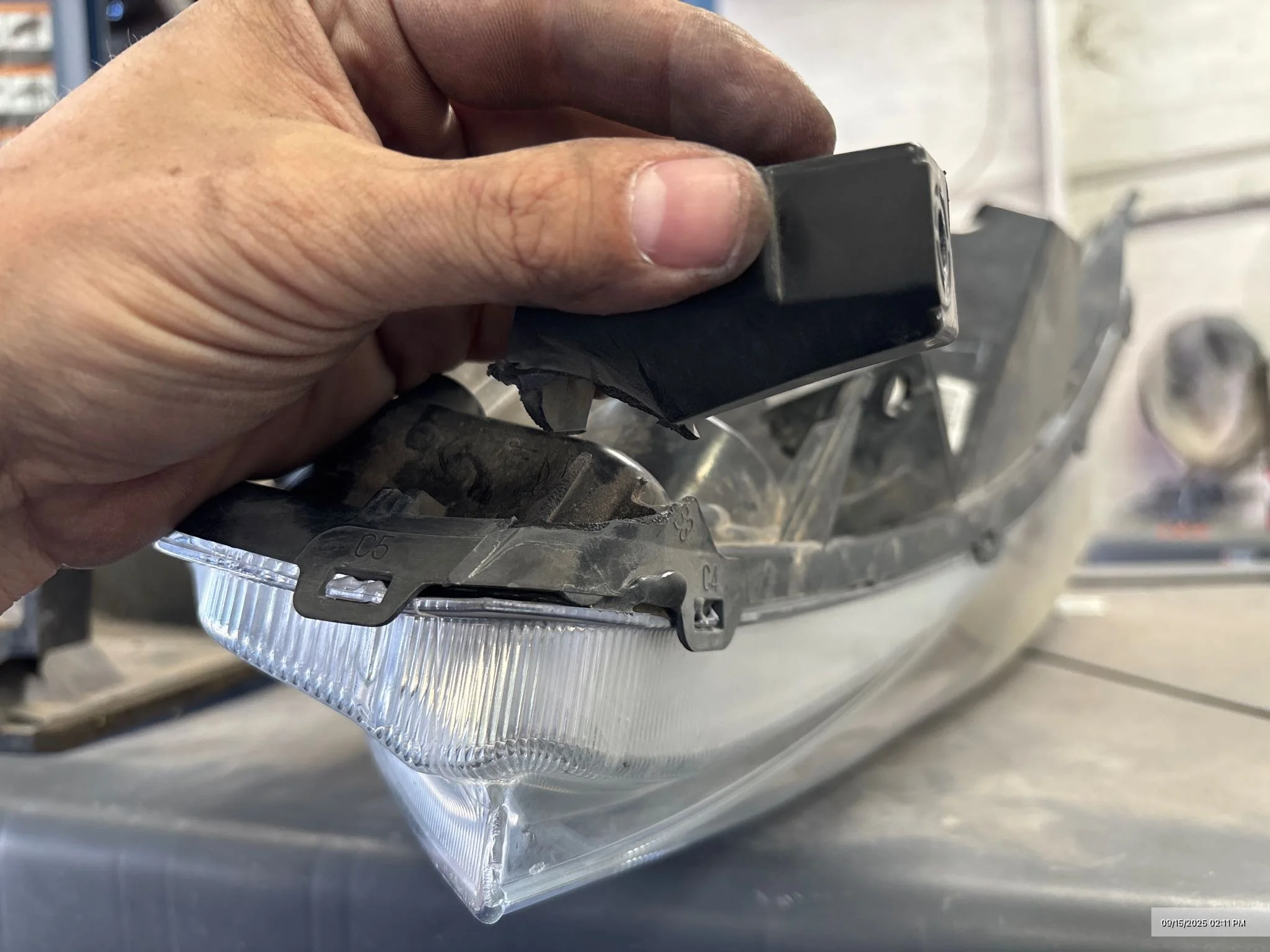 Close-up of a hand holding a damaged black plastic automotive headlight assembly, exposing the inner reflective surface and lens, with a blurred background of a workshop or garage.