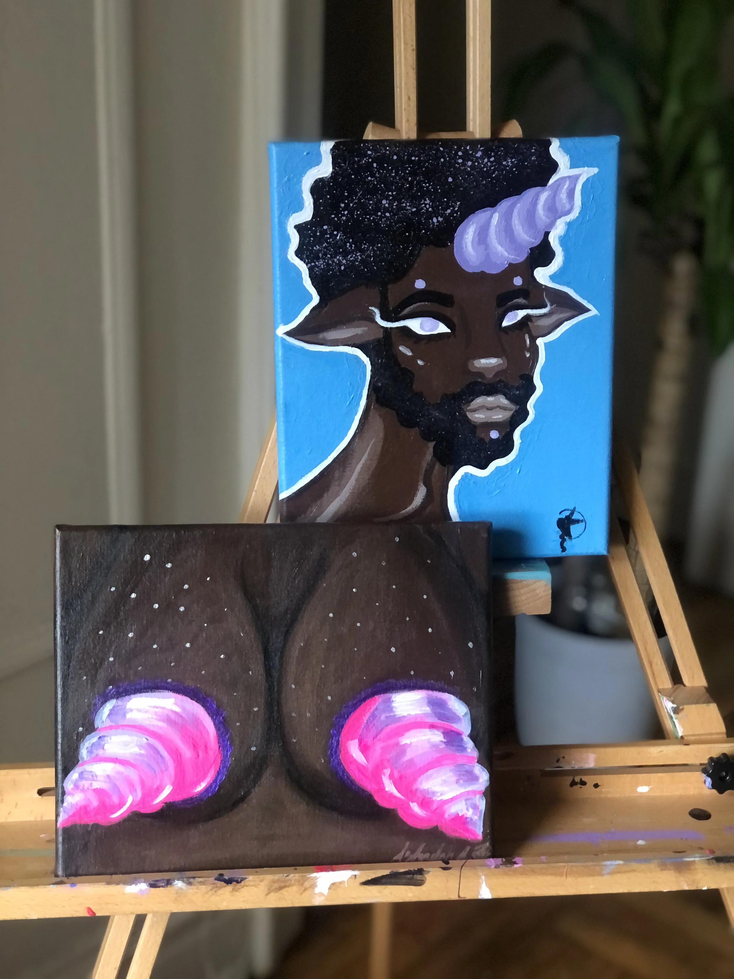 Two paintings on a wooden easel. The top painting is a portrait of a dark-skinned figure with a unicorn horn, starry hair, and white eyes with no pupils, against a blue background. The bottom painting is an abstract depiction of a woman's buttocks wi