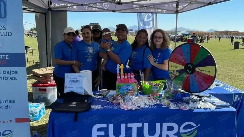 Grupo de personas en un puesto en un evento al aire libre, con una rueda de la fortuna y varias mesas con productos y regalos, todos vistiendo camisetas azules.