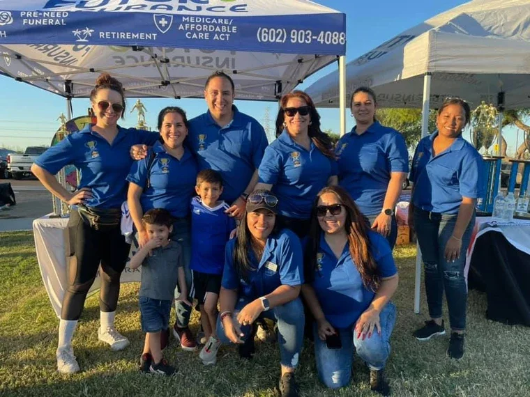 Grupo de ocho personas, adultos y niños, posando en un evento al aire libre con toldos azules y blancos, algunos sosteniendo boletos y objetos promocionales, todos con camisetas azules, parece un evento comunitario o de salud.