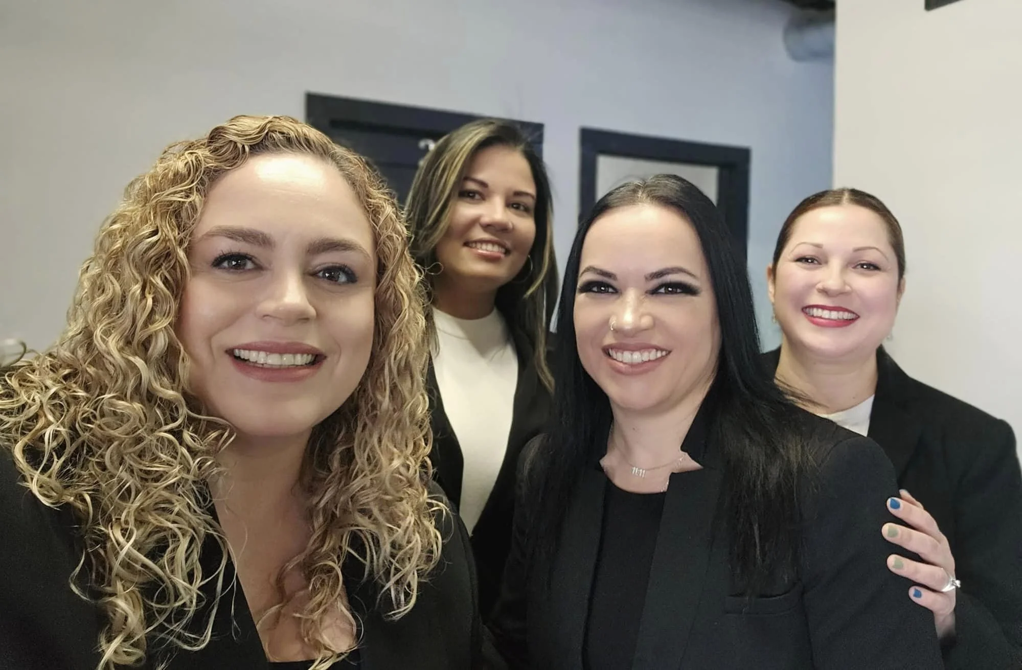Cuatro mujeres sonrientes en una reunión o evento, vestidas con ropa formal negra, en un entorno interior.