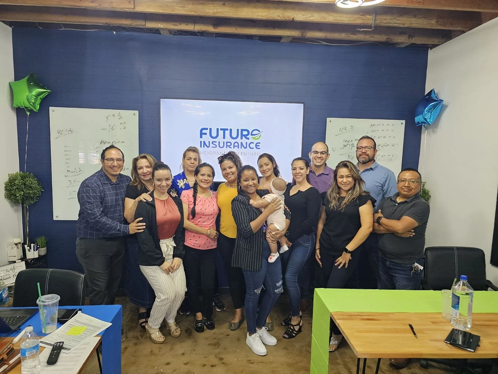 Grupo de personas en una reunión o celebración en una sala de oficina, con una pantalla que dice 'FUTURE INSURANCE' y decoraciones con globos.