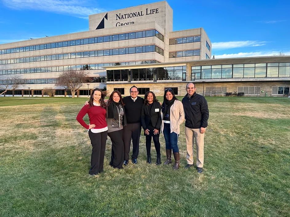 Grupo de seis personas sonriendo en frente de un edificio de oficinas con el letrero 'National Life Group', en un día soleado con cielo azul y pocas nubes, en un área de césped