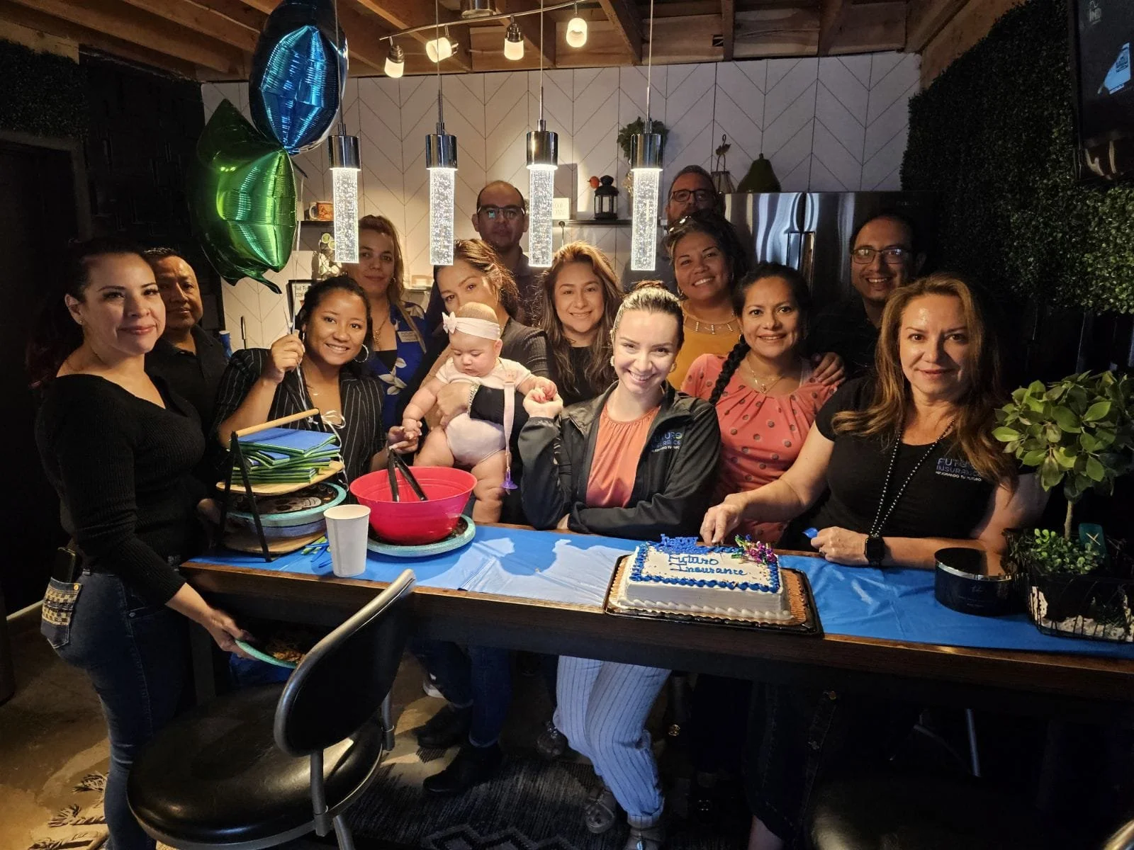 Un grupo de personas celebrando un cumpleaños en un restaurante, con un pastel en la mesa y globos. Todos sonrientes, algunos de pie y otros sentados, con decoración festiva en el fondo.