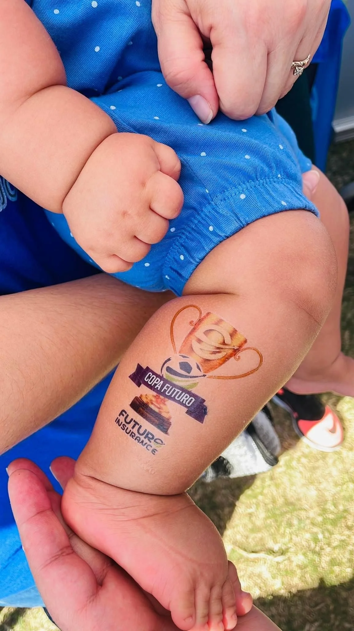 Un bebé con la mano cerrada en forma de puño, usando un short azul con puntos blancos, mostrando en su pierna un tatuaje temporal del logotipo de la Copa Fútbol y seguro de futuras aseguradoras.