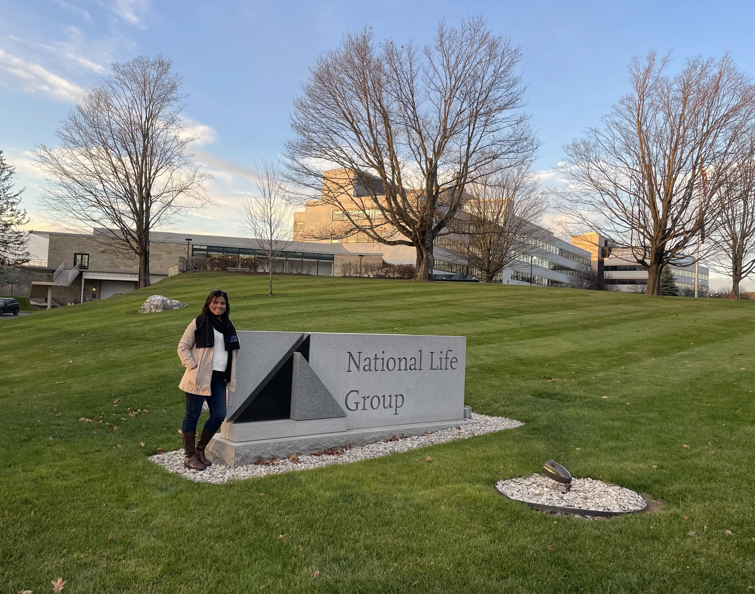 Mujer parada junto a un cartel de piedra que dice 'National Life Group' en un terreno con césped y árboles sin hojas, con un edificio moderno de fondo bajo un cielo despejado.