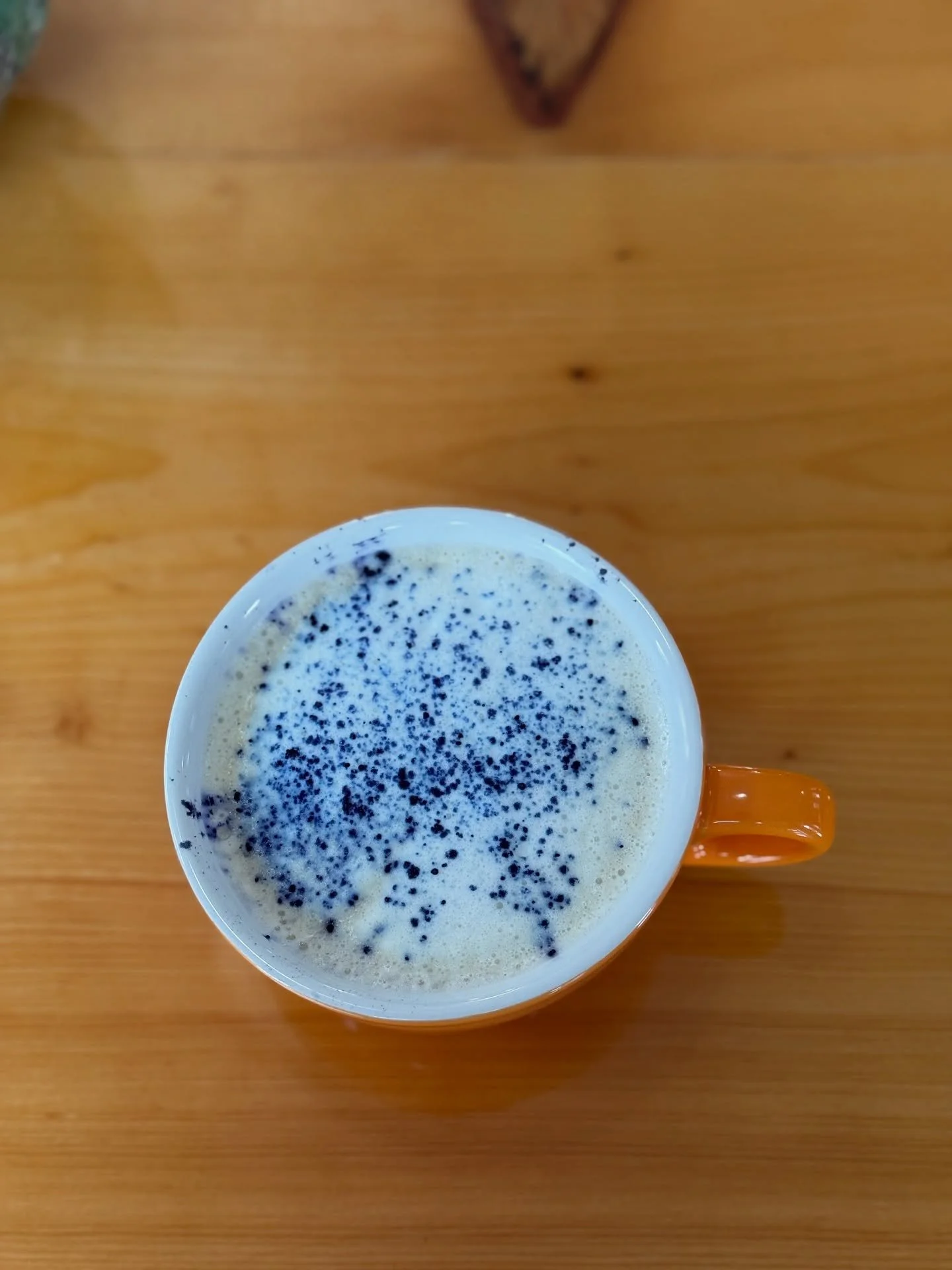 Top view of a white mug with orange handle filled with a foamy drink topped with butterfly pea flower specks, placed on a wooden surface.