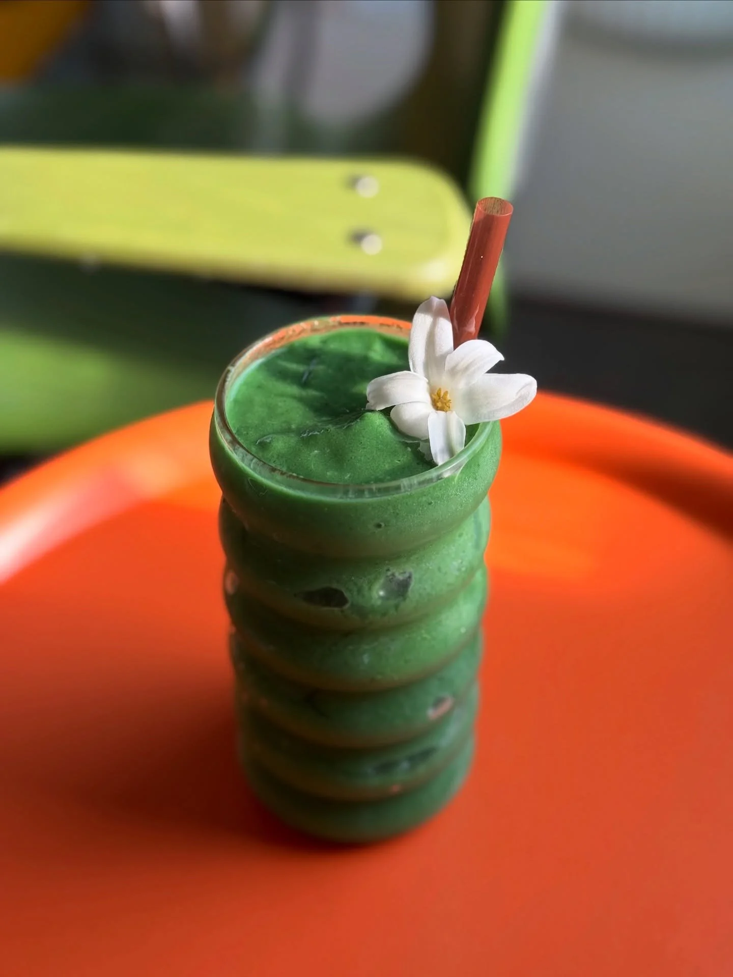 Green layered smoothie in a textured glass with a white flower and red straw, on an orange surface with a green background.