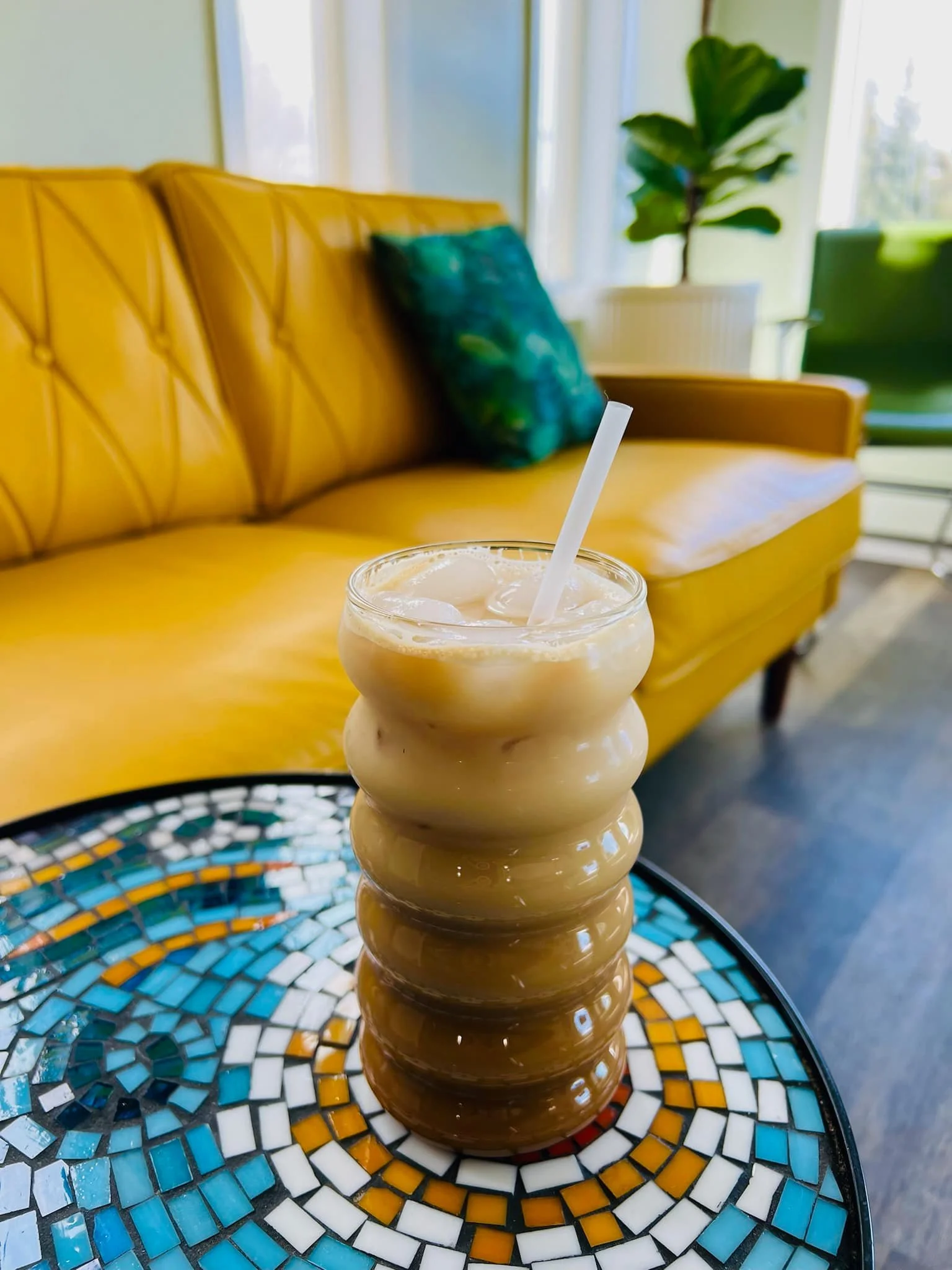 A tall, multi-layered iced coffee or smoothie in a ribbed glass with a straw on a colorful mosaic table in a bright, modern cafe. In the background, a yellow sofa with a green pillow, a large leafy plant, and windows with white curtains.