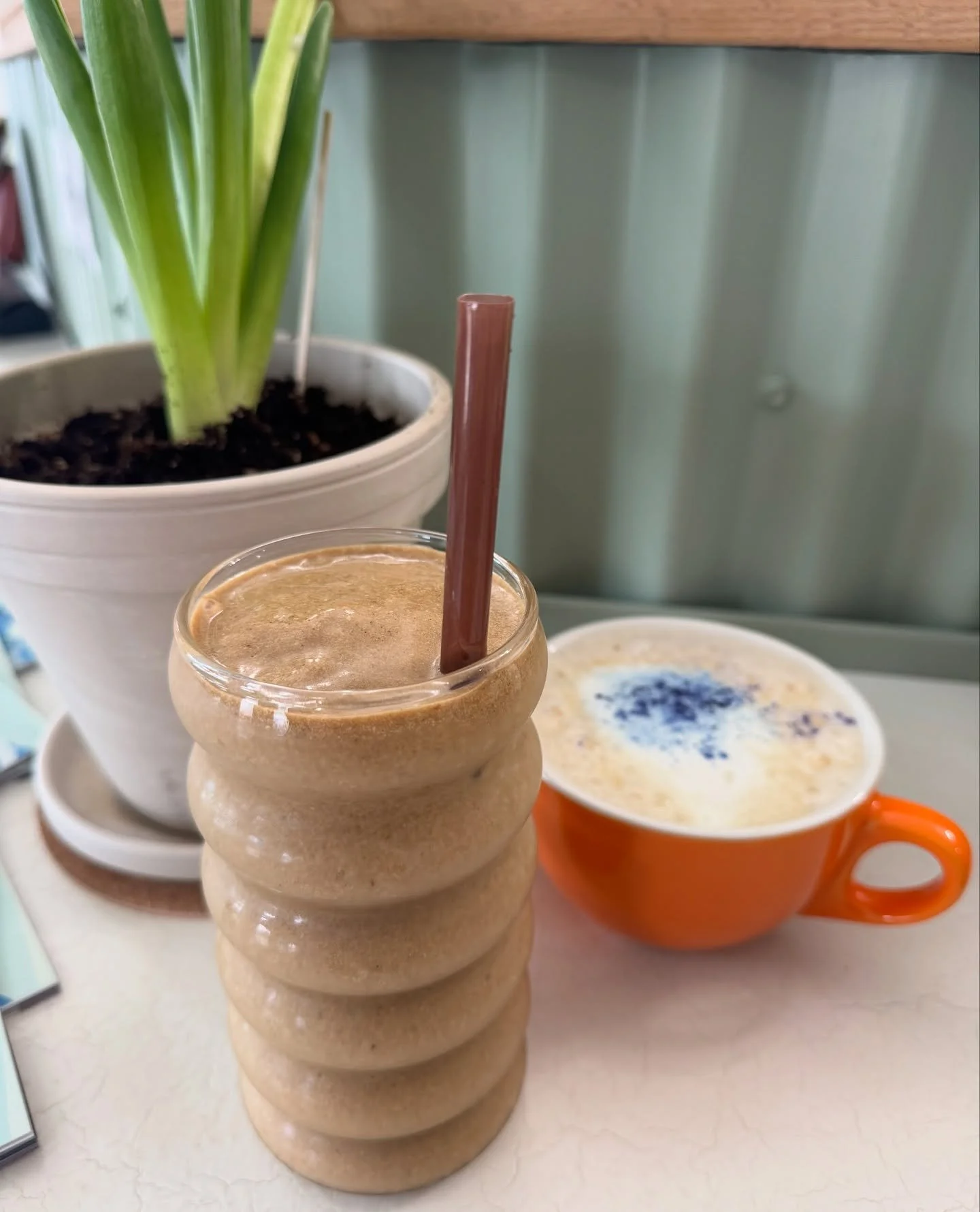 A layered glass filled with a blended coffee drink with a brown straw, an orange mug with a frothy beverage topped with blue sprinkles, and a potted plant with green leaves in the background on a light-colored table.