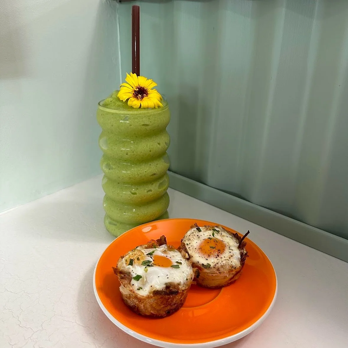 A green smoothie with a yellow flower and a red straw in a tall, wavy glass, placed against a light-colored wall. In front, on an orange plate, two eggs baked in bacon cups topped with herbs.