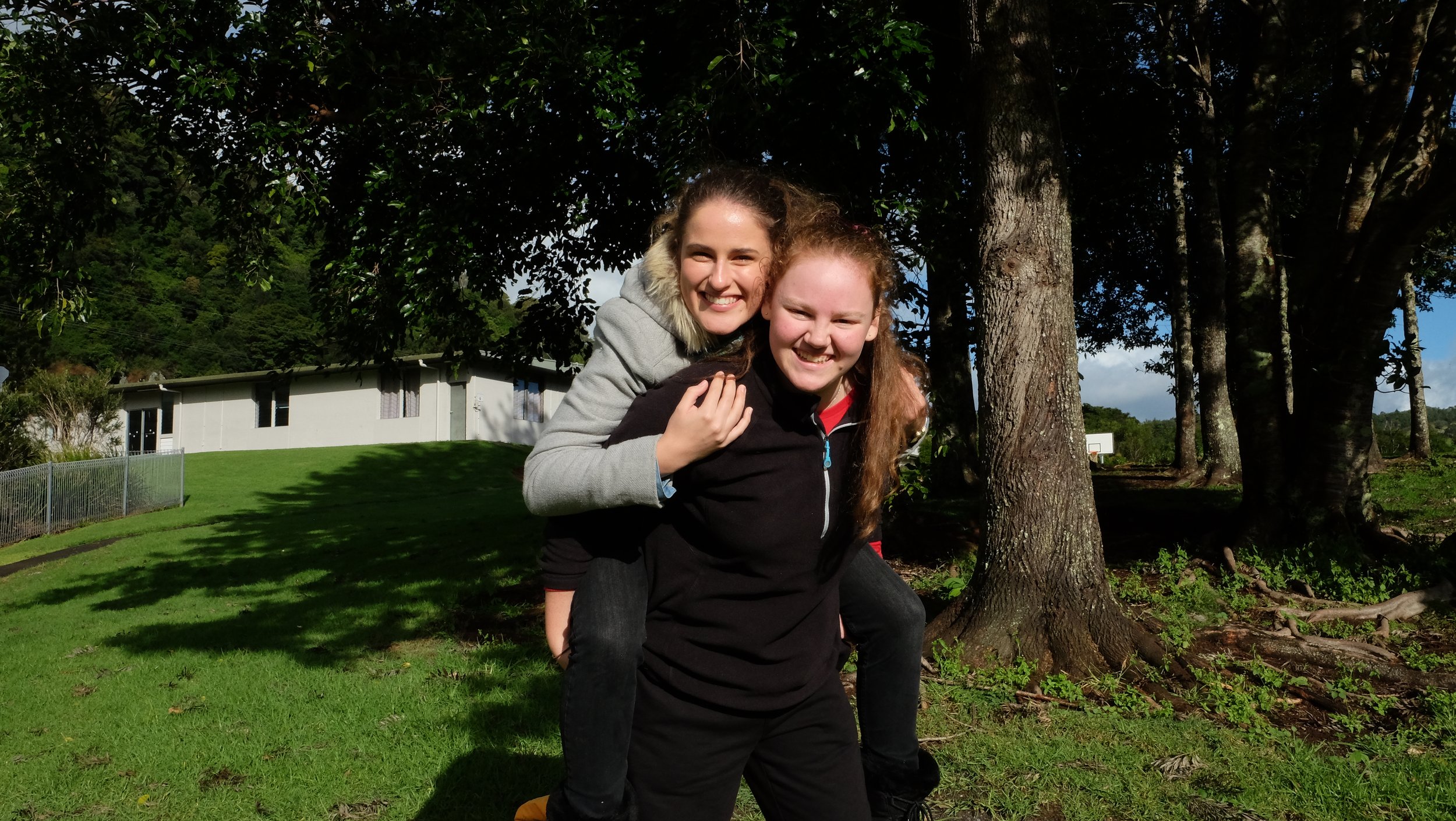 Two young women smile and pose outdoors in front of tall trees, with a house and blue sky in the background on a sunny day.