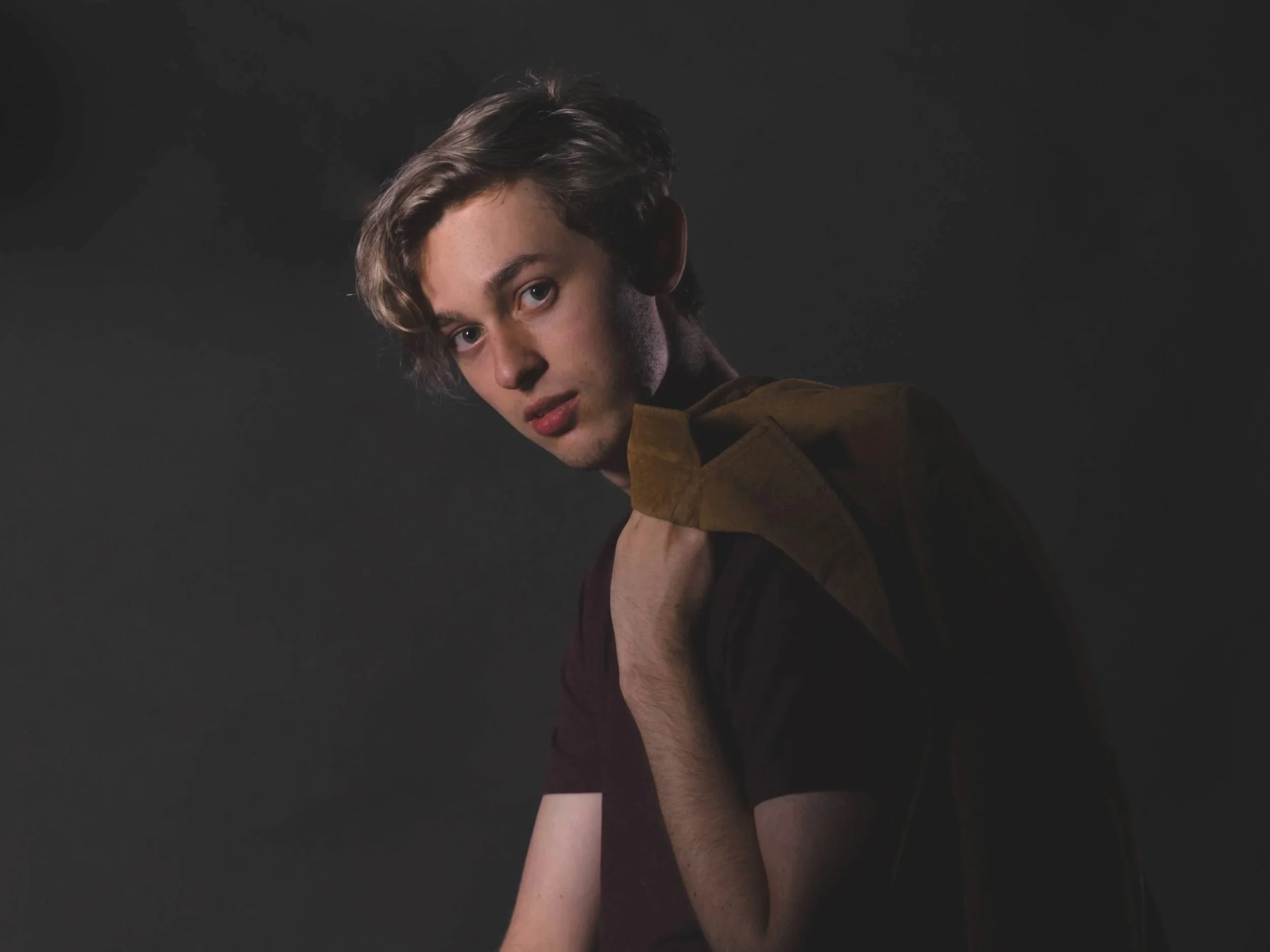 A young man with light skin, tousled brown hair, and wearing a brown jacket, looking at the camera with a neutral expression, against a dark background.