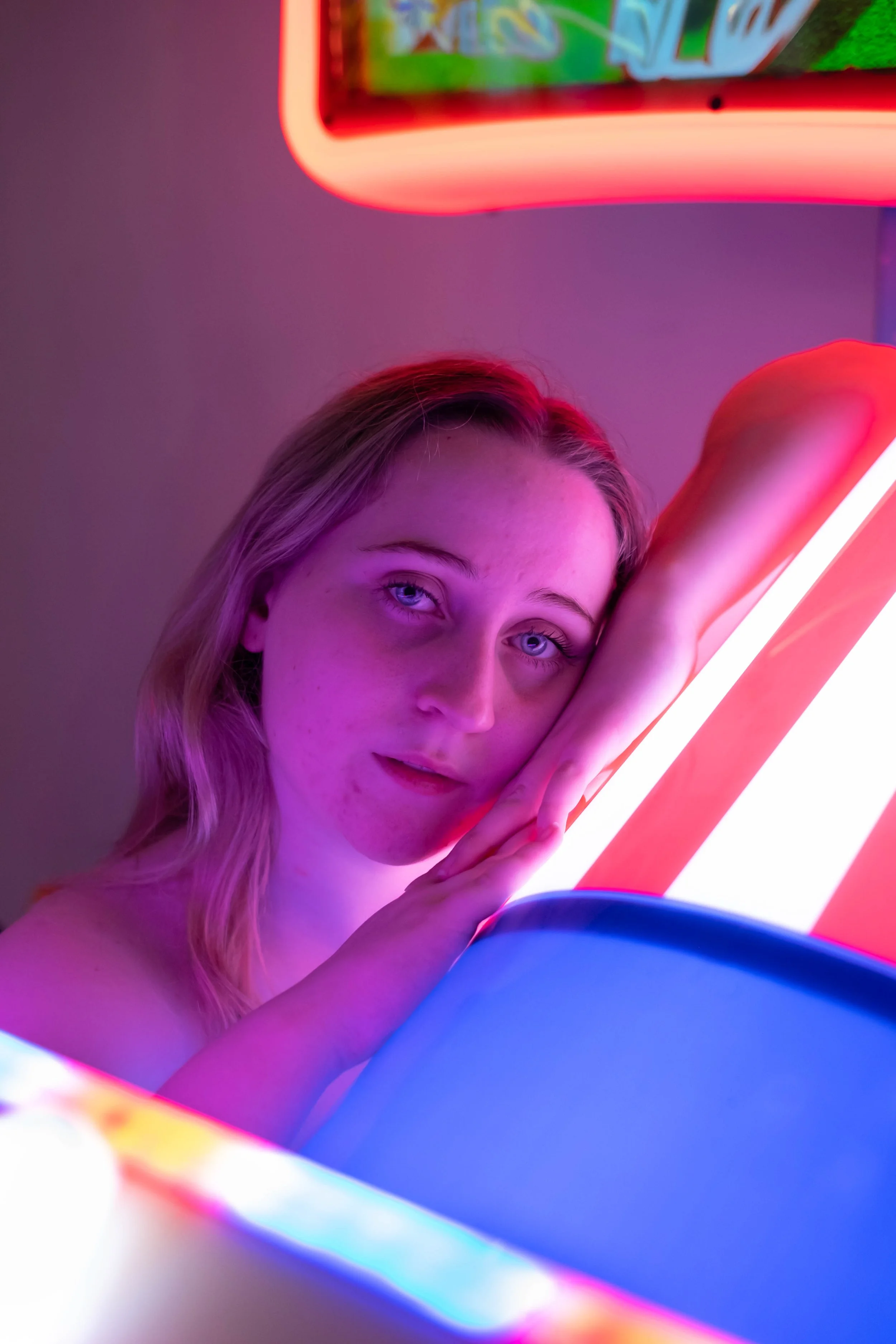 A young woman with blonde hair and blue eyes is illuminated by neon lights, creating a vibrant, colorful atmosphere. She rests her head on her hand, gazing at the camera with an expressionless face, surrounded by pink, purple, and blue neon glow.