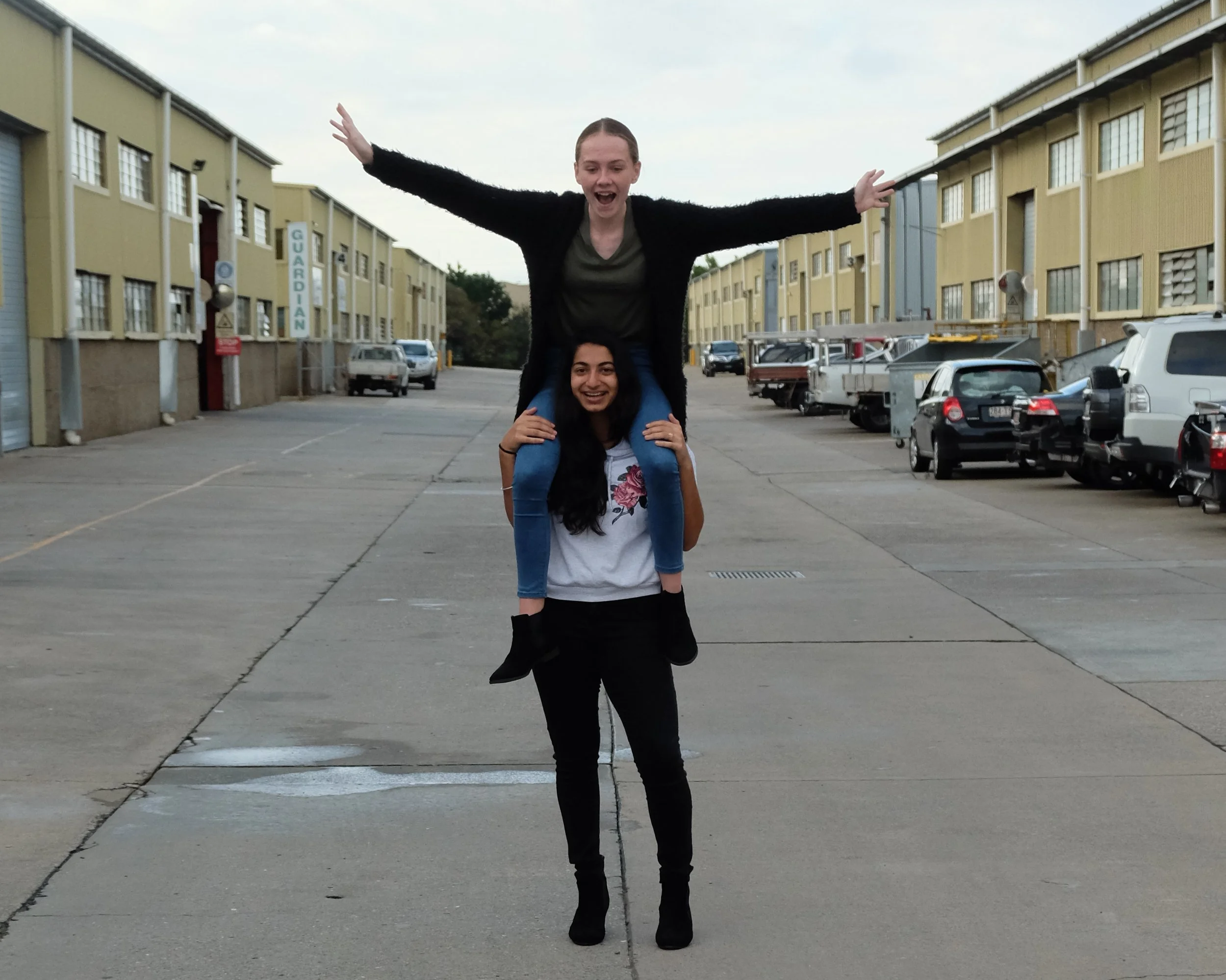 Two young women are outdoors in an industrial parking lot, with one woman sitting on the other's shoulders. The woman on top has her arms outstretched and is smiling, while the woman on the bottom is smiling as well.