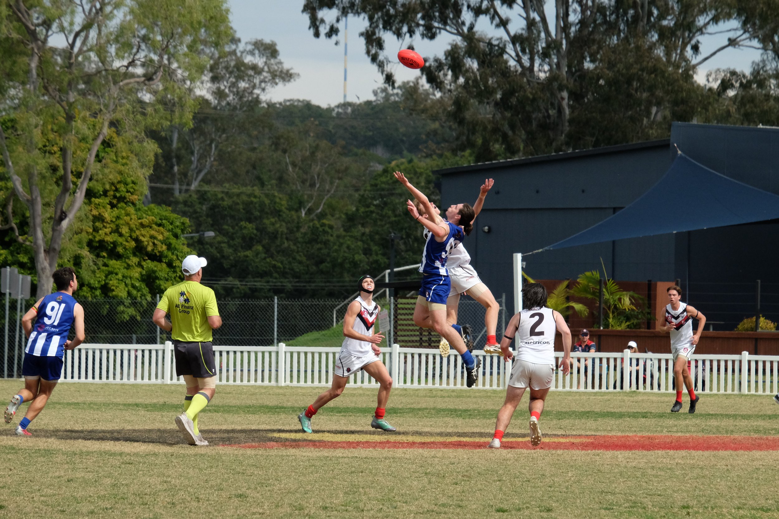 A group of people playing Australian Rules football outdoors on a grassy field, with some players jumping to catch the ball and others watching or moving nearby, surrounded by trees.