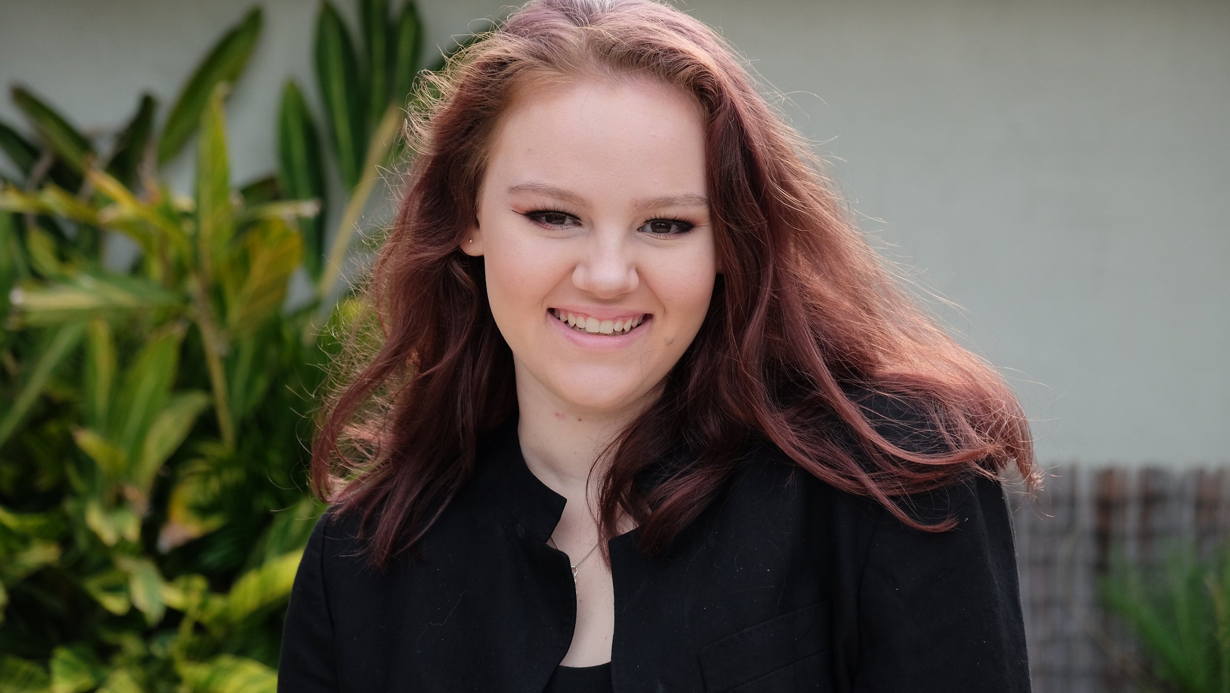 A woman with red hair smiling outdoors, wearing a black top, with green foliage in the background.