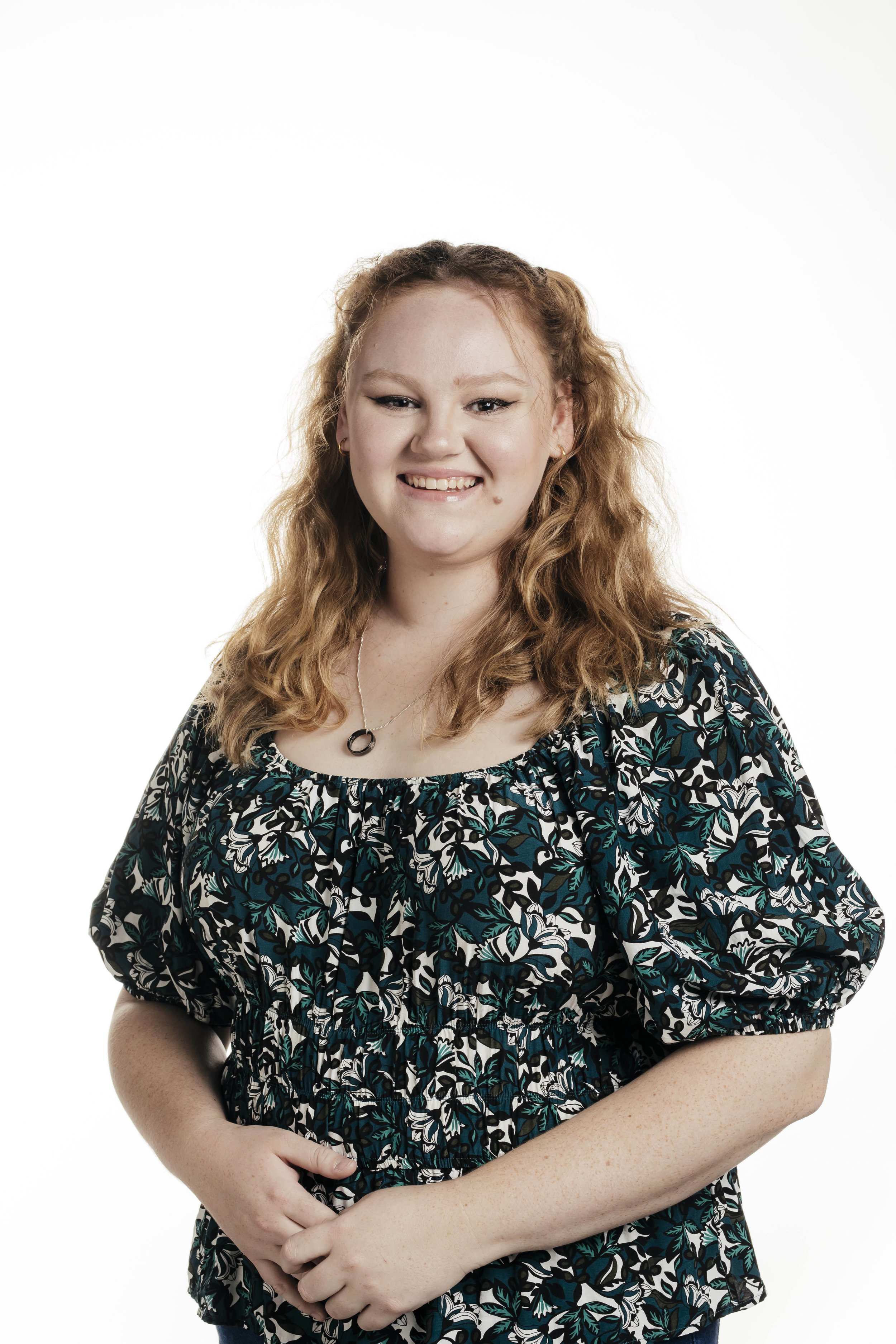 Young woman with curly red hair smiling, wearing a patterned dress and jewelry, standing against a white background.