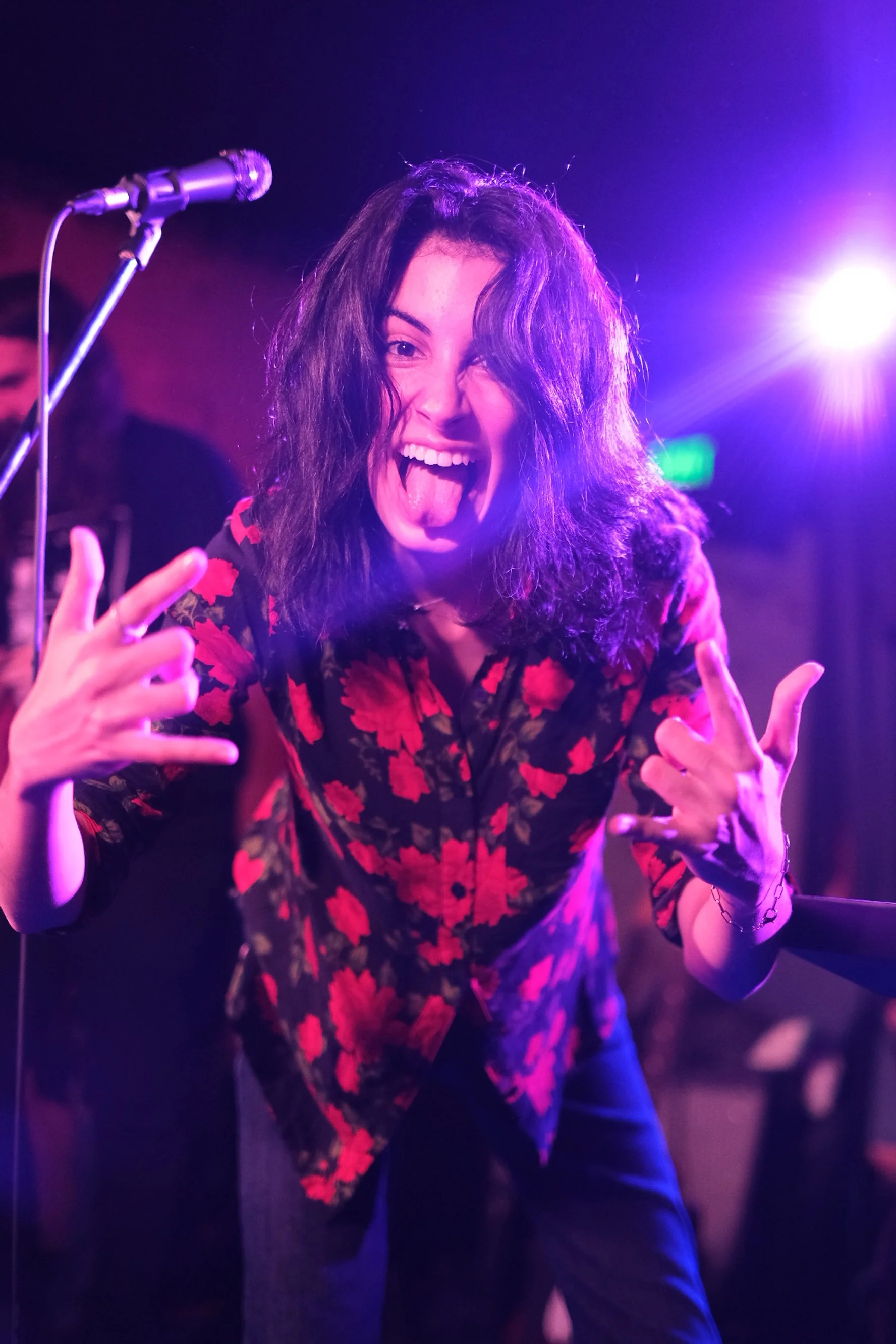 A woman with curly dark hair making a rock sign with one hand and sticking her tongue out in a lively, colorful setting with purple and pink lighting.