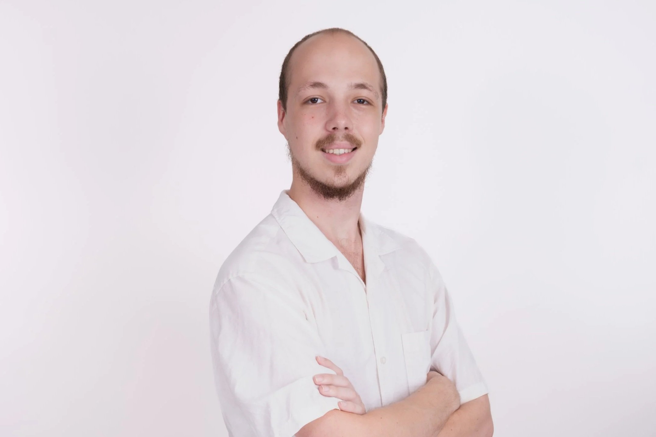 A young man with a goatee and short hair, wearing a white shirt, stands with arms crossed against a white background, smiling