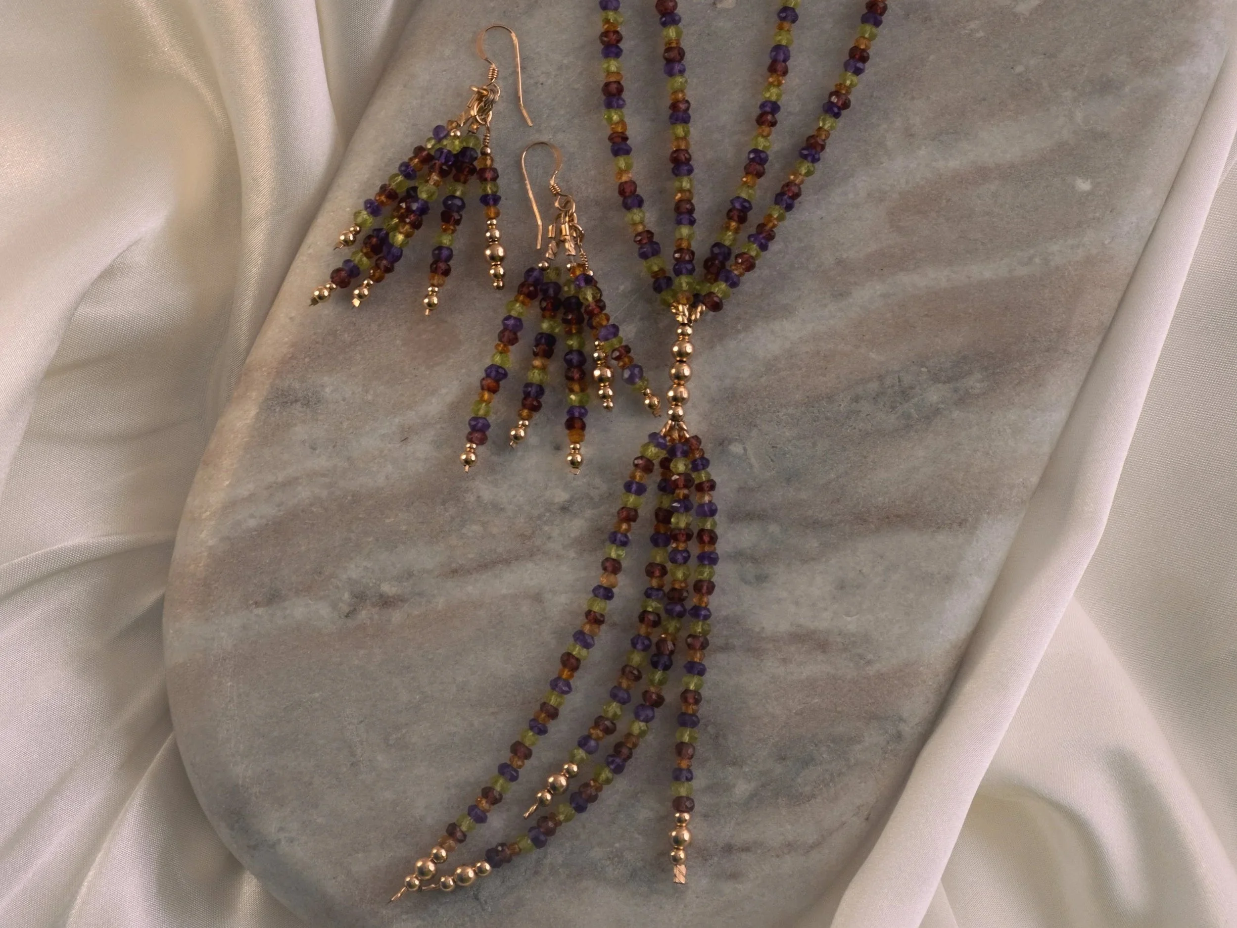 Colorful beaded jewelry set including necklaces and earrings with purple, green, red, gold, and transparent beads, displayed on a gray surface with white fabric background.