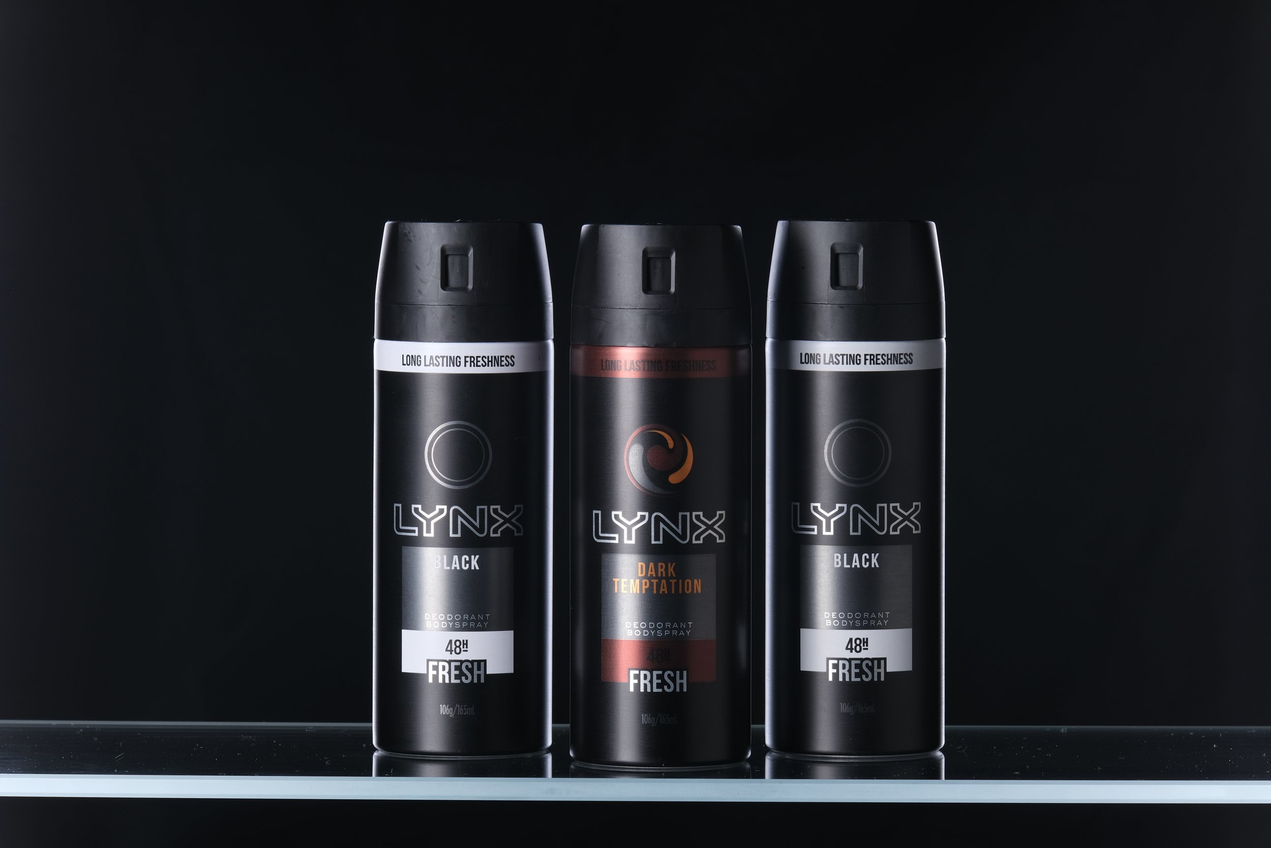 Three black cans of Lynx body spray on a dark background, with the middle one labeled 'Dark Temptation' and the other two labeled 'Black,' featuring a minimalist design and the phrase 'Long Lasting Freshness'.