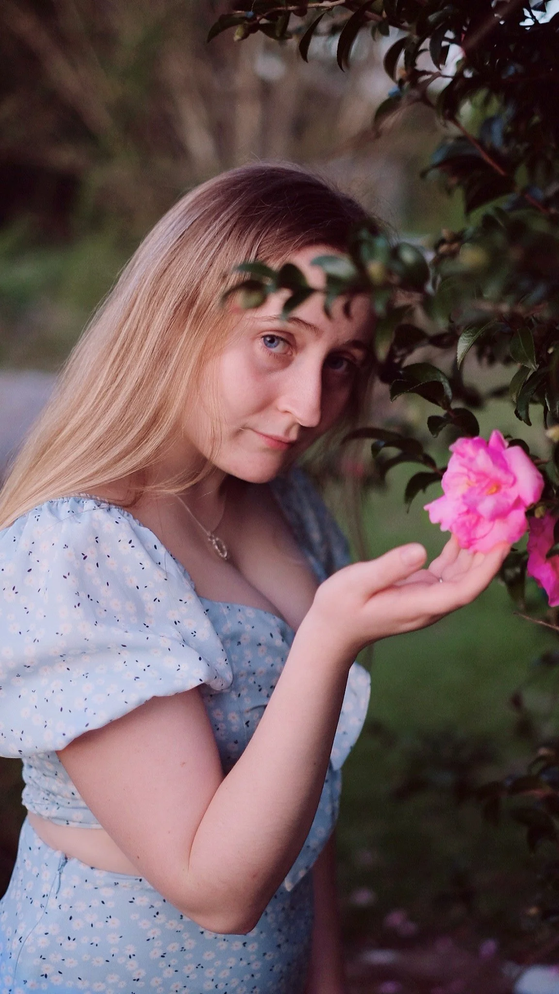 A young woman with long blonde hair and blue eyes holding a pink flower near bushes with green leaves.