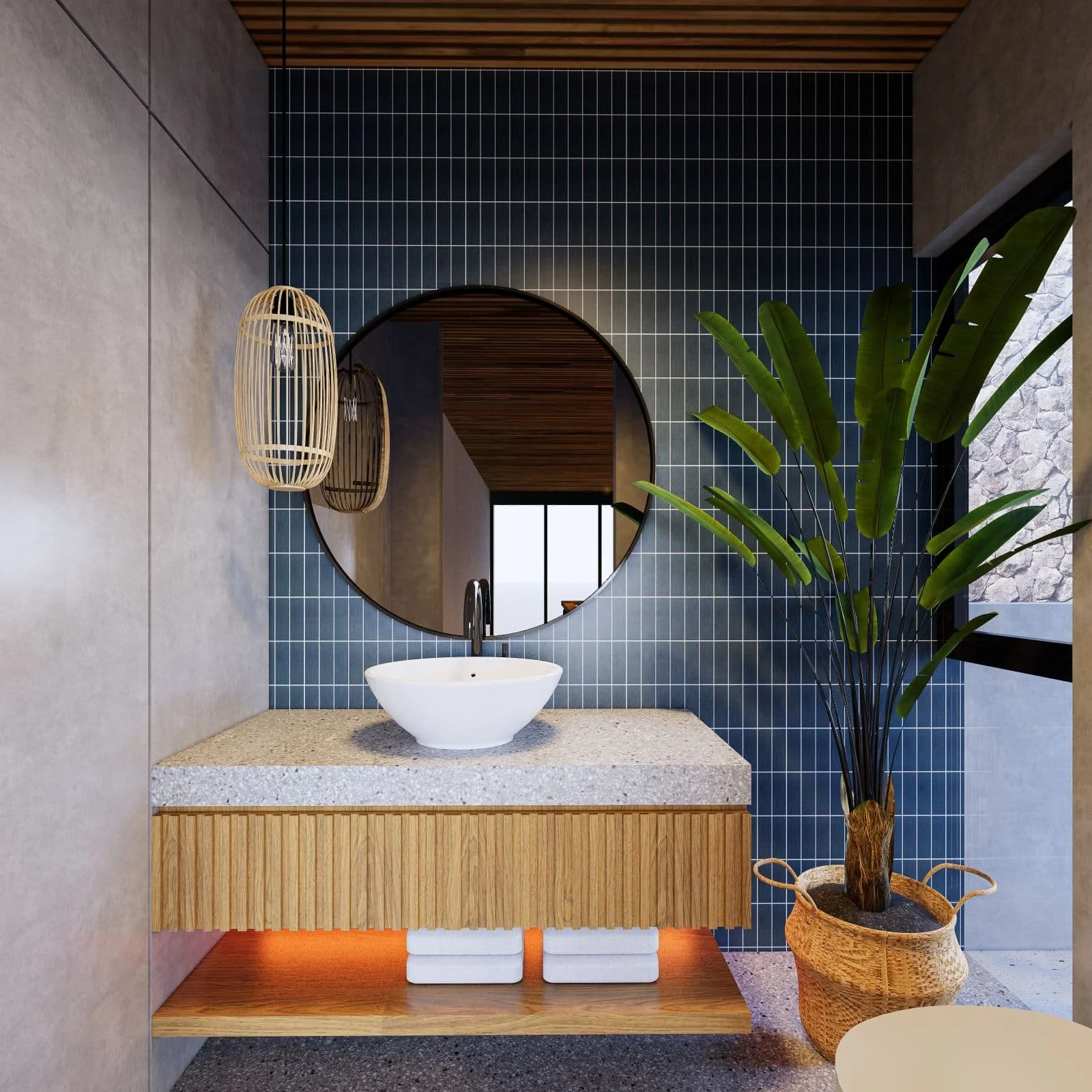 A modern bathroom with a round mirror above a white vessel sink on a speckled gray countertop. The wall behind the mirror is decorated with small dark blue tiles, and to the right, there is a large potted plant with broad green leaves. Two wicker pen