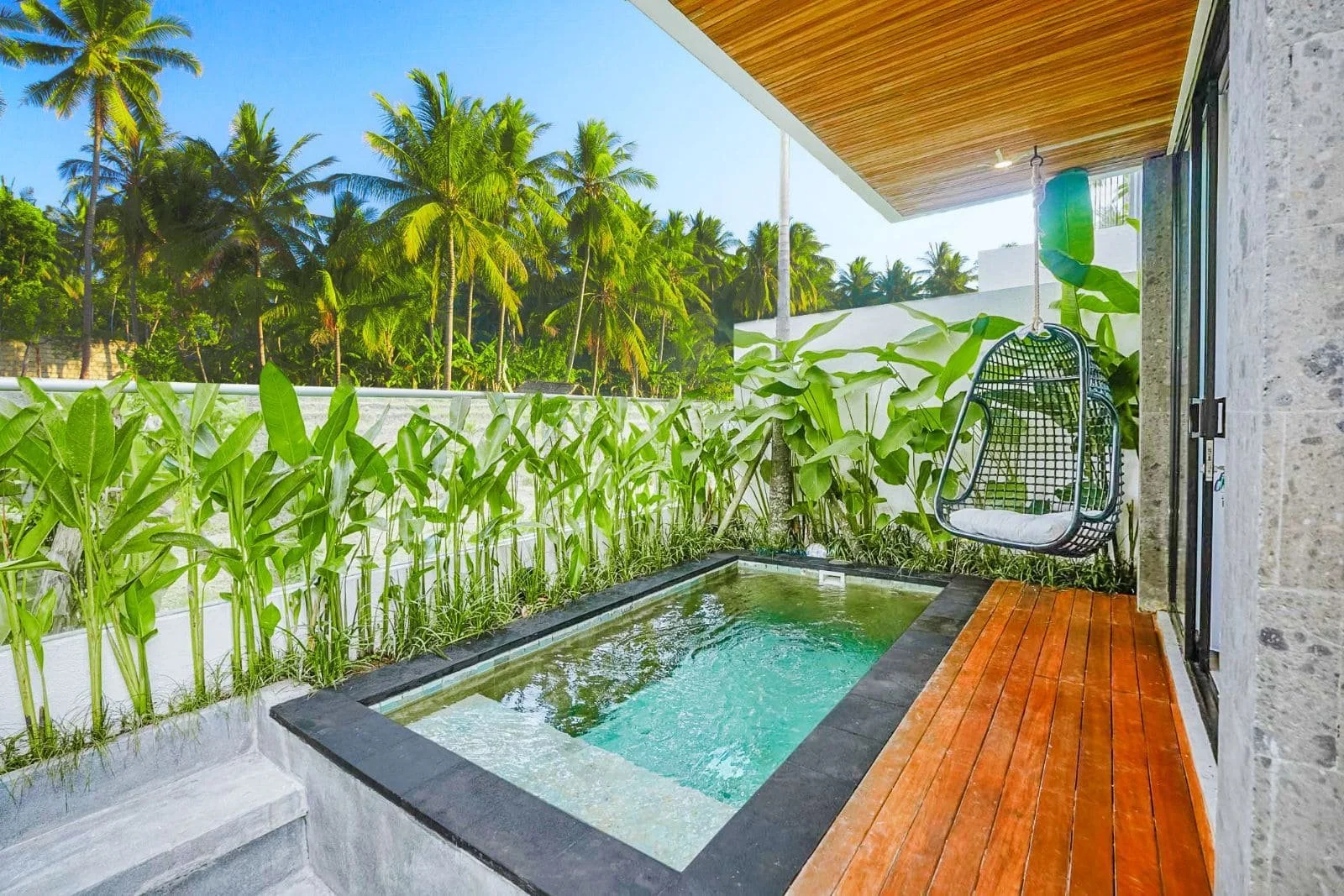 Small outdoor patio with a built-in hot tub, surrounded by green plants and trees, with a wooden deck and a hanging black patio swing.