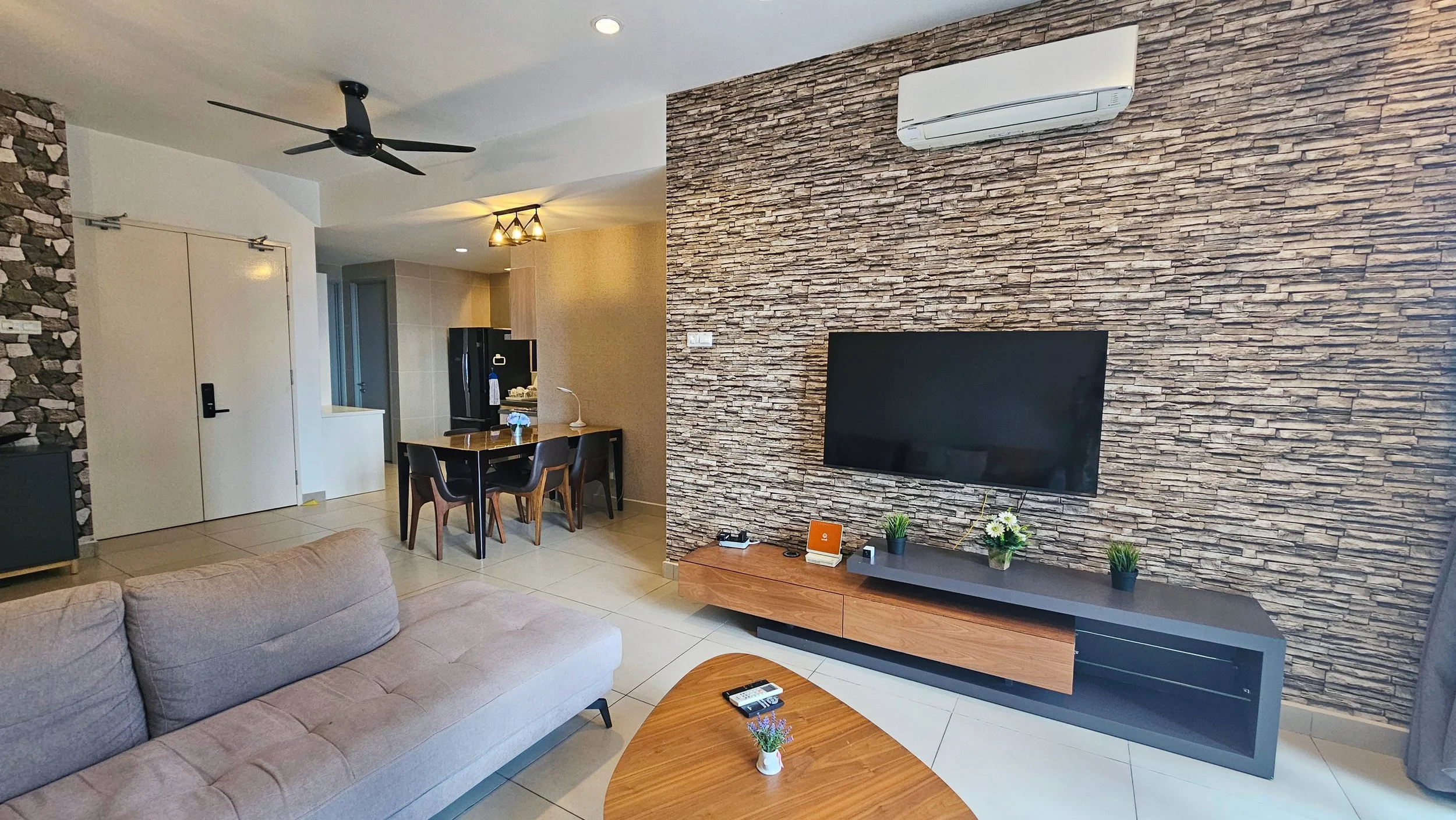 Modern living room with a beige sofa, a wooden coffee table, a TV mounted on a stone accent wall, and a media console with plants. Kitchen and dining area visible in the background with a black table, chairs, and a refrigerator.