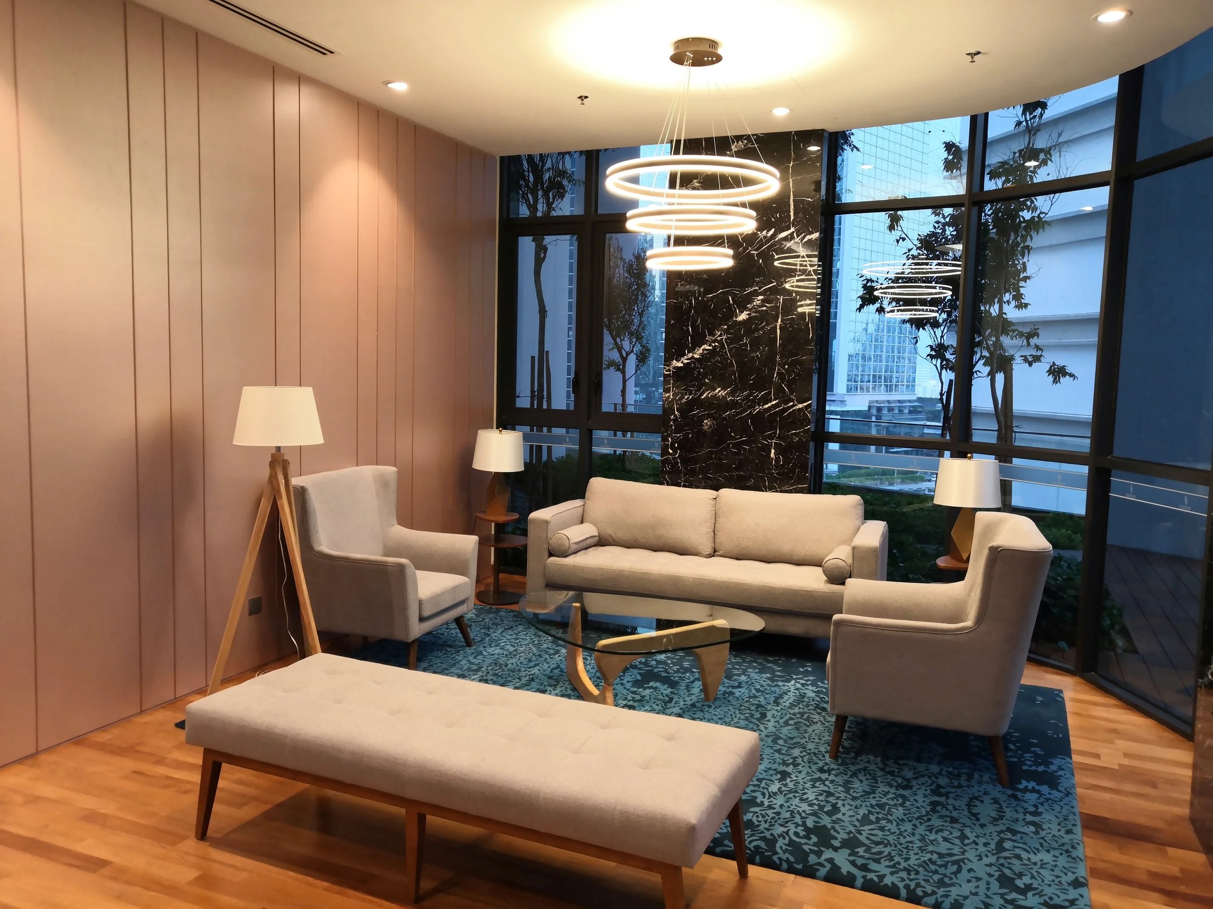 Elegant living room with beige upholstered sofas, armchairs, a glass coffee table, a tufted ottoman, and wooden side tables with lamps, large floor-to-ceiling windows, modern circular pendant lights, and cityscape view outside.