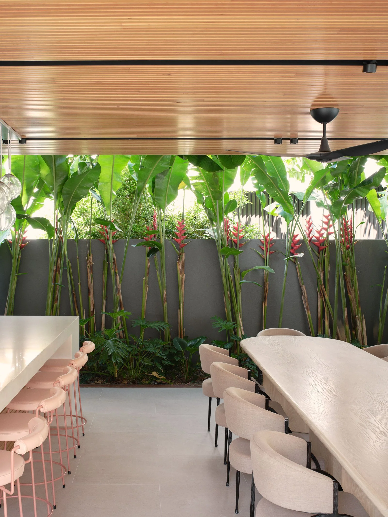 Interior of a modern dining area with a long light wood table surrounded by beige chairs. Behind the table, there is a lush green wall of large leafy plants and red ginger flowers, with a natural light source coming through the greenery. The ceiling