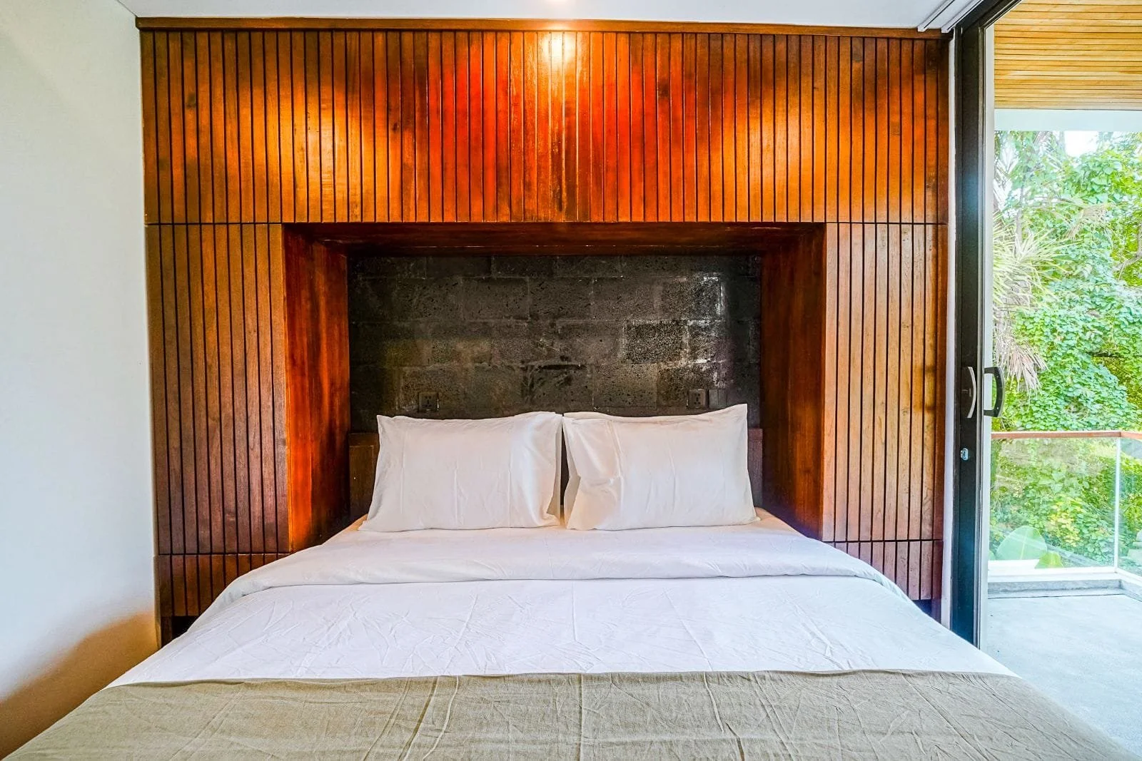 A bed with white bedding and two pillows positioned against a dark stone headboard, surrounded by a wooden frame with vertical slats, next to a glass sliding door opening to an outdoor balcony with greenery.