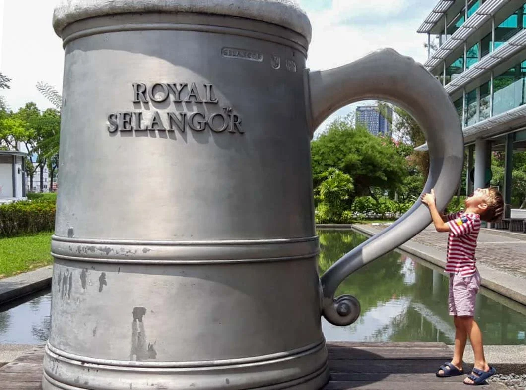 A child standing next to a large metallic mug sculpture with 'Royal Selangor' inscription, outdoors in a park-like setting.