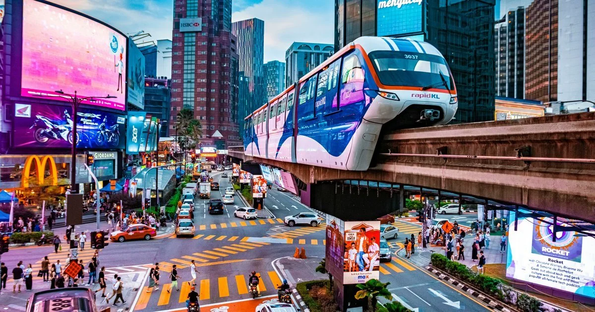 Bright city scene in an urban area during the day with a monorail train traveling on an elevated track above busy streets filled with cars and pedestrians. Tall buildings with digital billboards and advertisements surround the intersection.