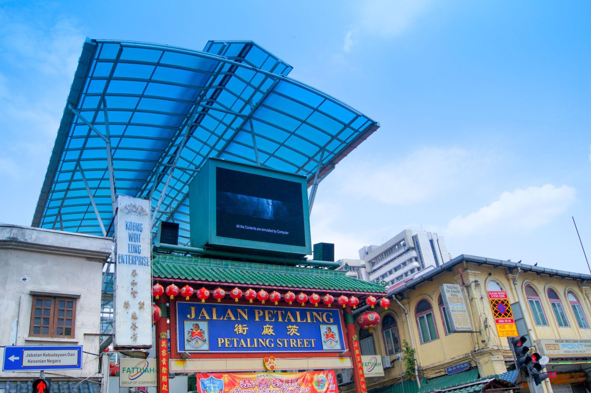Street scene with traffic lights, signs, and building facades, including a prominent sign that reads 'Jalan Petaling' and another banner with Chinese characters, under a blue sky.