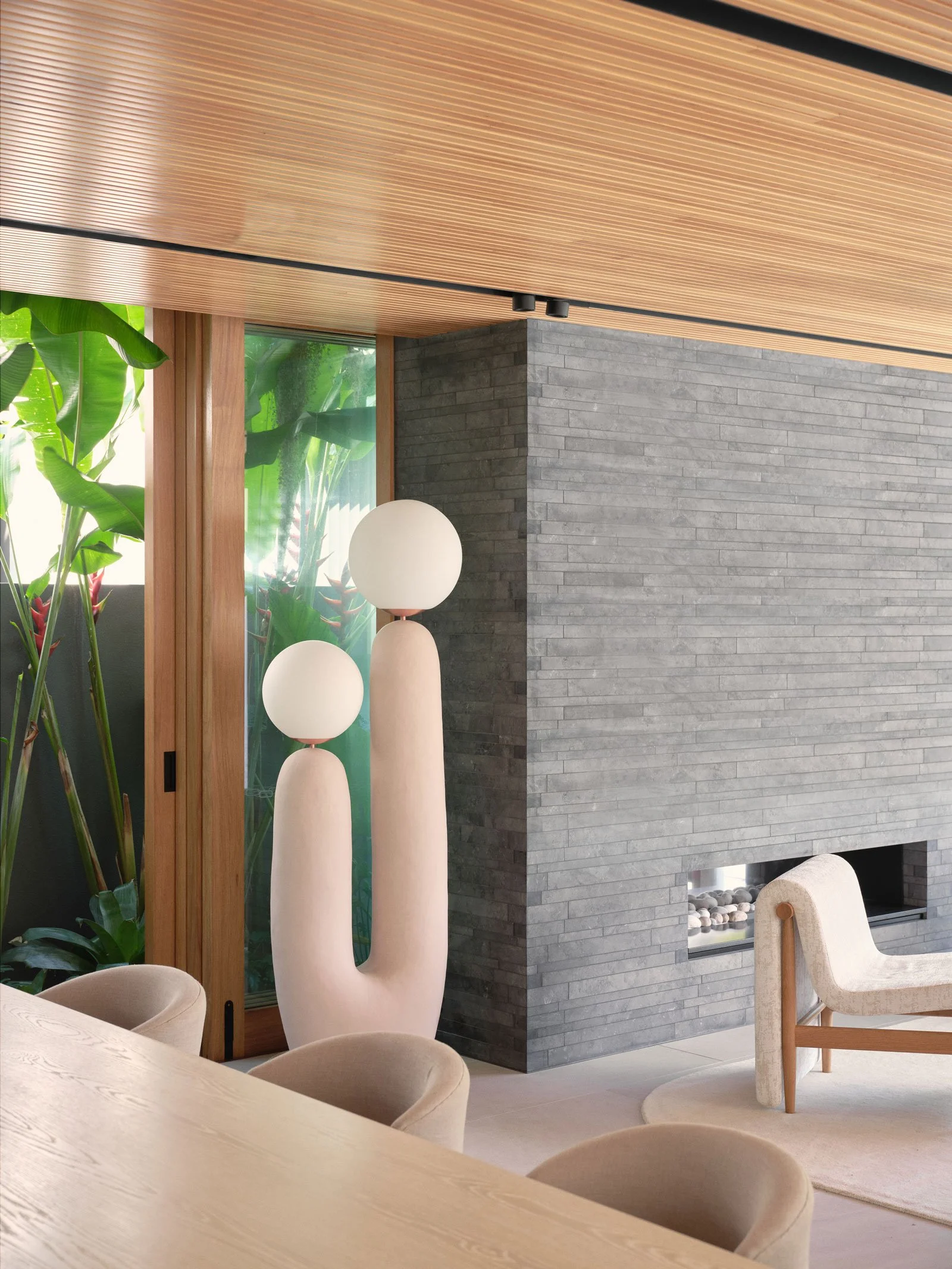 Modern interior living space featuring a black brick fireplace, wooden ceiling, large windows with green plants outside, and a decorative white sculpture with spherical lamps on top, along with beige upholstered chairs and a light wood table.