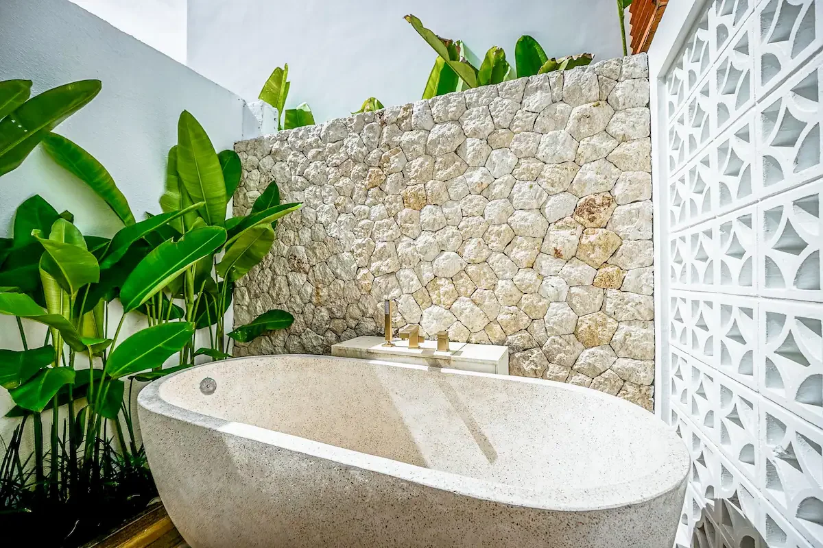 An outdoor bathroom with a freestanding bathtub, tropical green plants, a stone wall, and a decorative white concrete block wall.