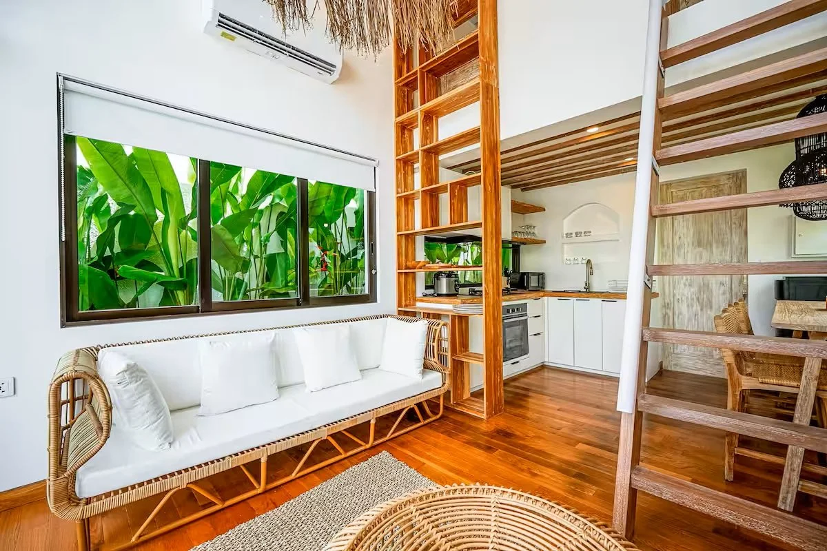 Interior of a modern tropical-style home featuring a white sofa, wooden flooring, and a large window with lush green leaves visible outside. The space includes an open kitchen with white cabinets, wooden shelves, and a staircase leading to an upper l