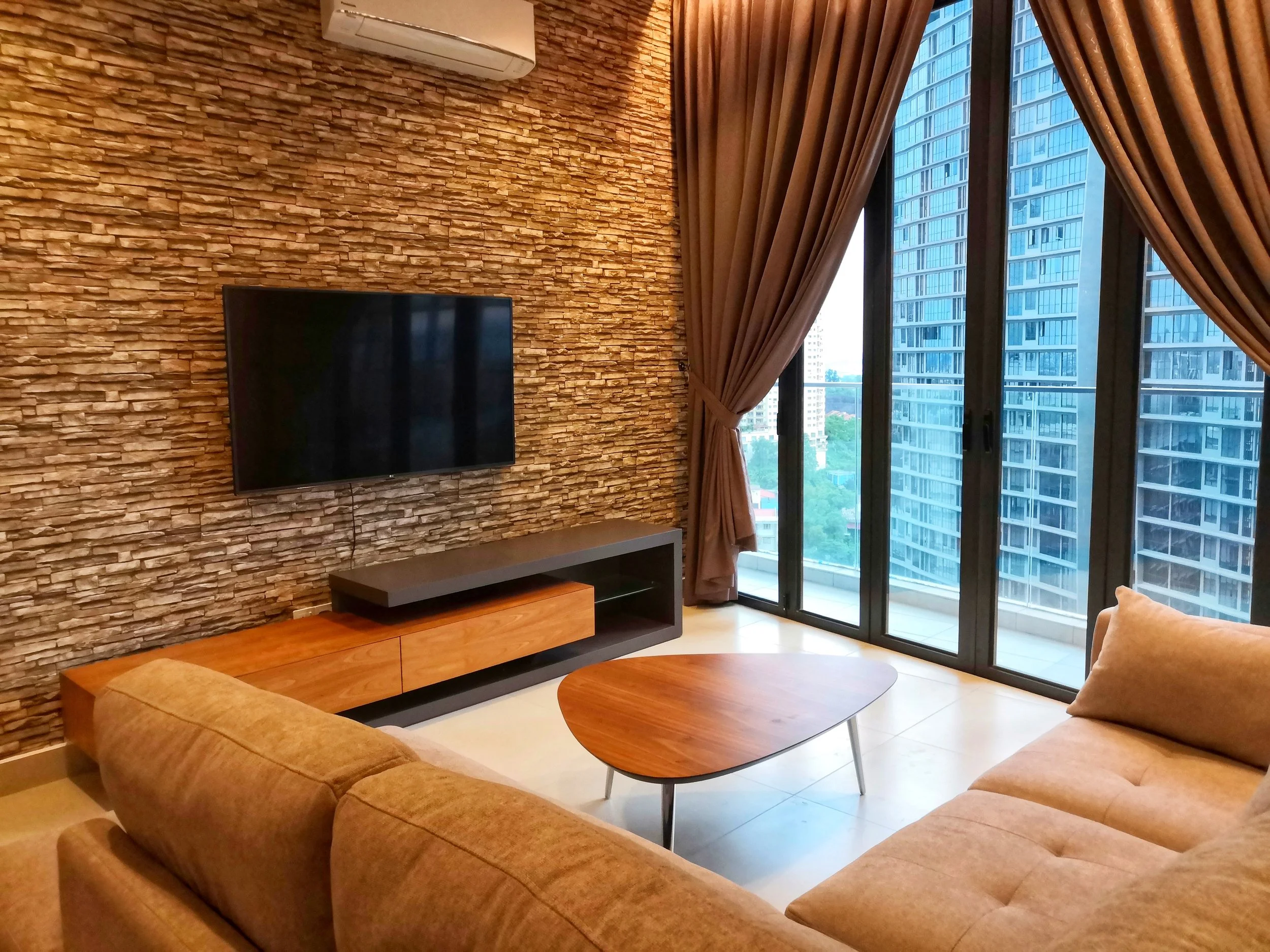 Modern living room with stone accent wall, wall-mounted flat-screen tv, beige sectional sofa, wooden coffee table, large windows with brown curtains, and city high-rise buildings visible outside.