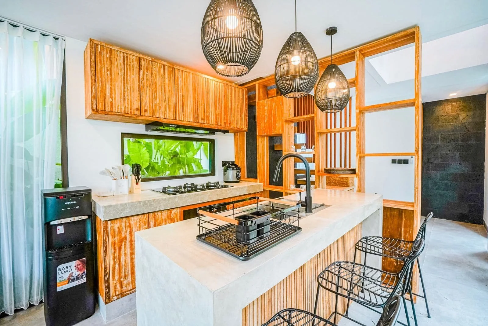 Modern kitchen with wooden cabinets, a concrete countertop, black bar stools, and hanging pendant lights. A water dispenser is next to a window with curtains.