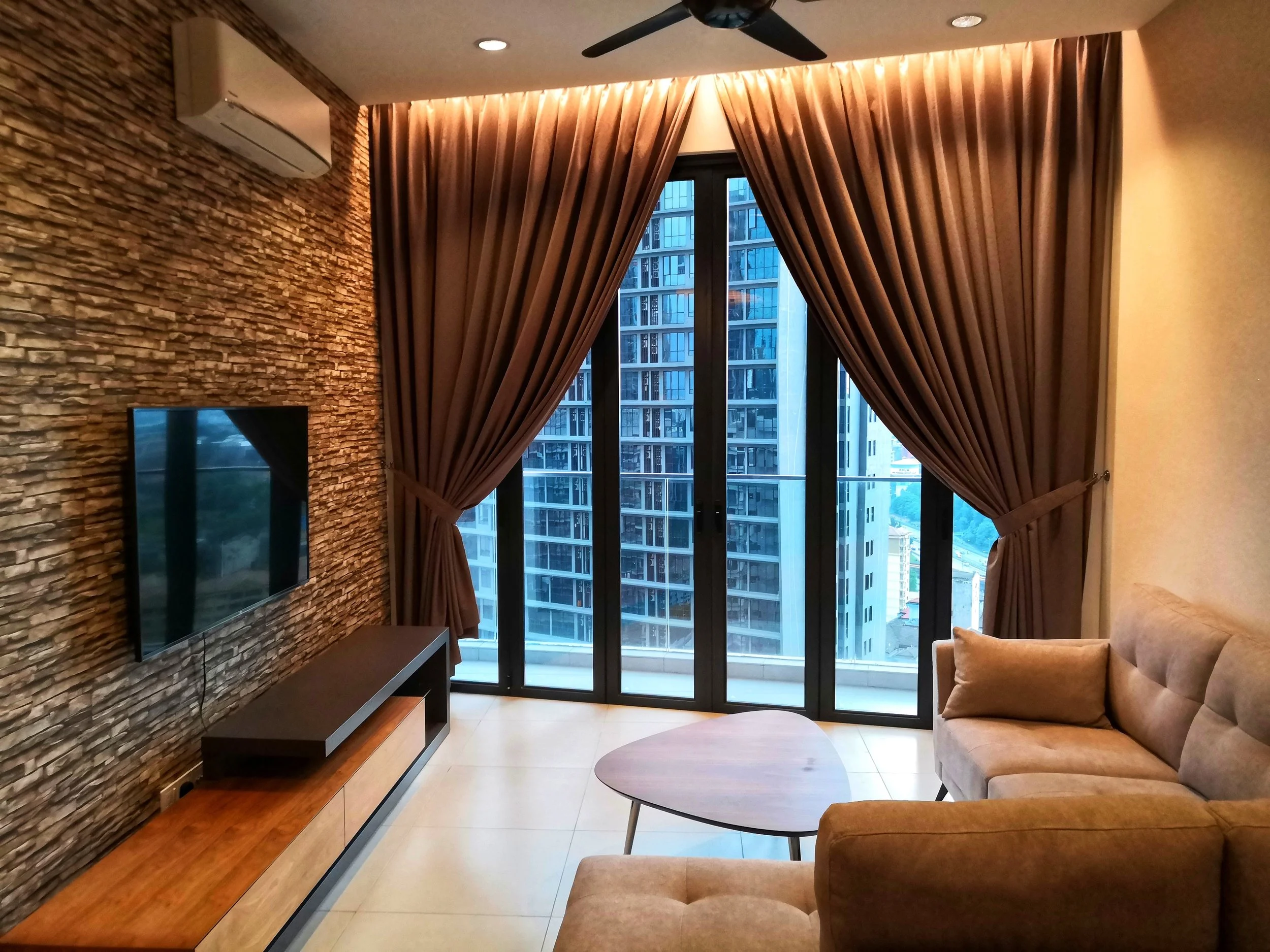 Living room with brick accent wall, mounted TV, wooden TV stand, beige sofa, round coffee table, large window with brown curtains, cityscape view, ceiling fan, wall-mounted air conditioner, beige tiled floor.