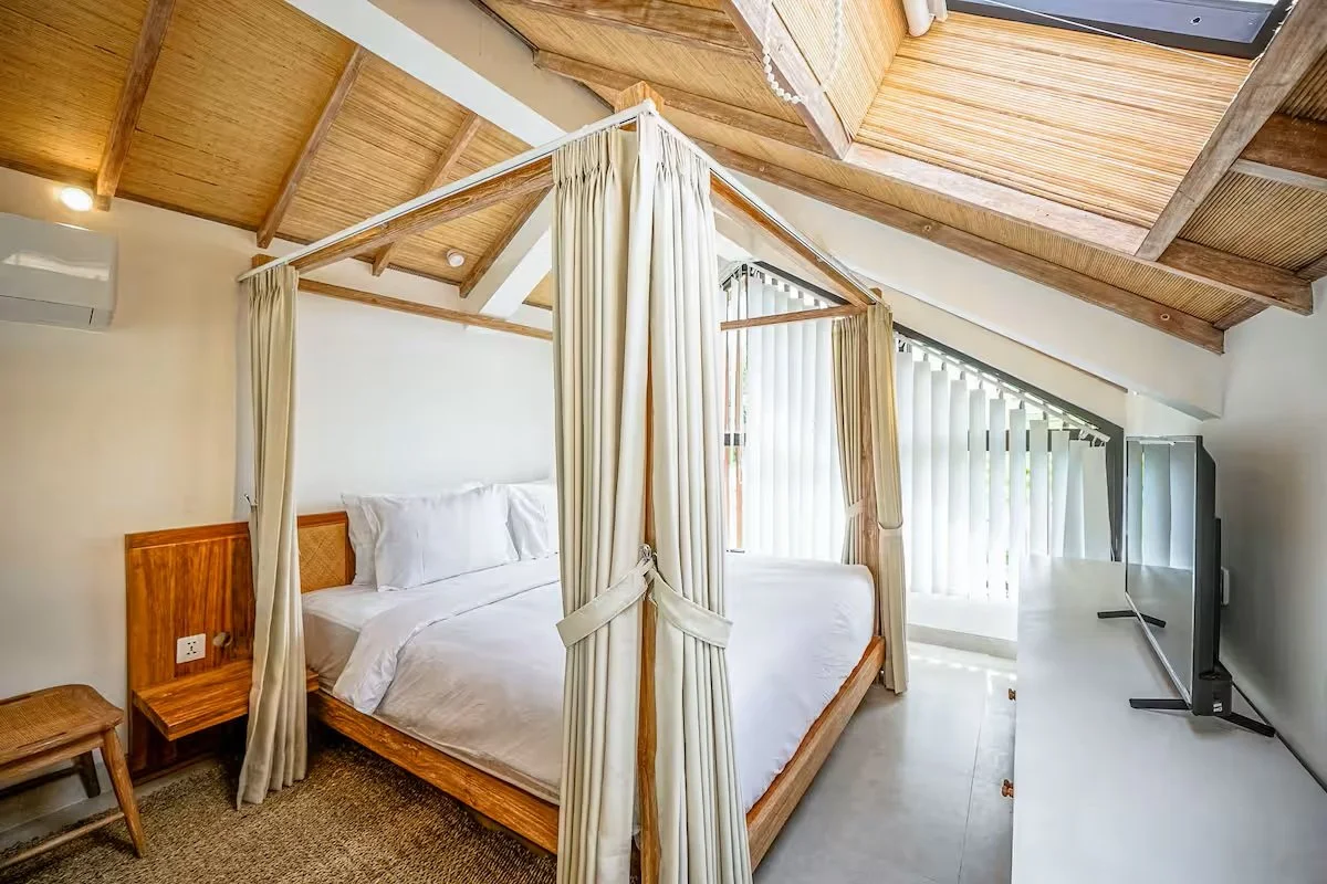 A cozy bedroom with a four-poster bed, cream curtains, white bedding, wooden furniture, vertical blinds, sloped wooden ceiling, and a flat-screen TV.