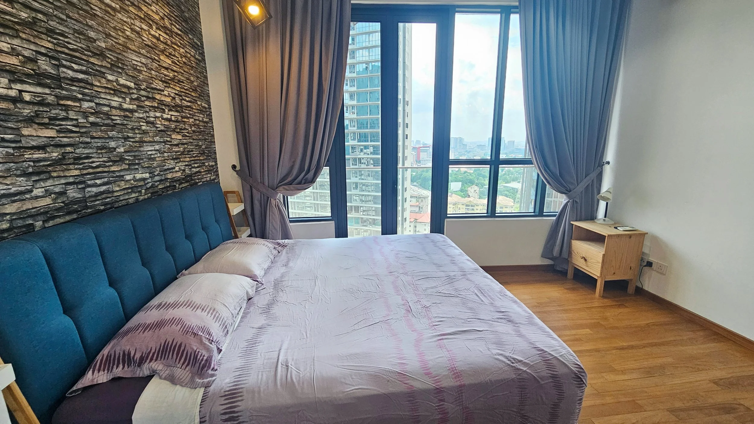 A bedroom with a large window showing city buildings, a bed with purple sheets and pillows, a stone accent wall behind the bed, metal curtain rods with purple curtains tied back, a wooden nightstand with a lamp and a phone.