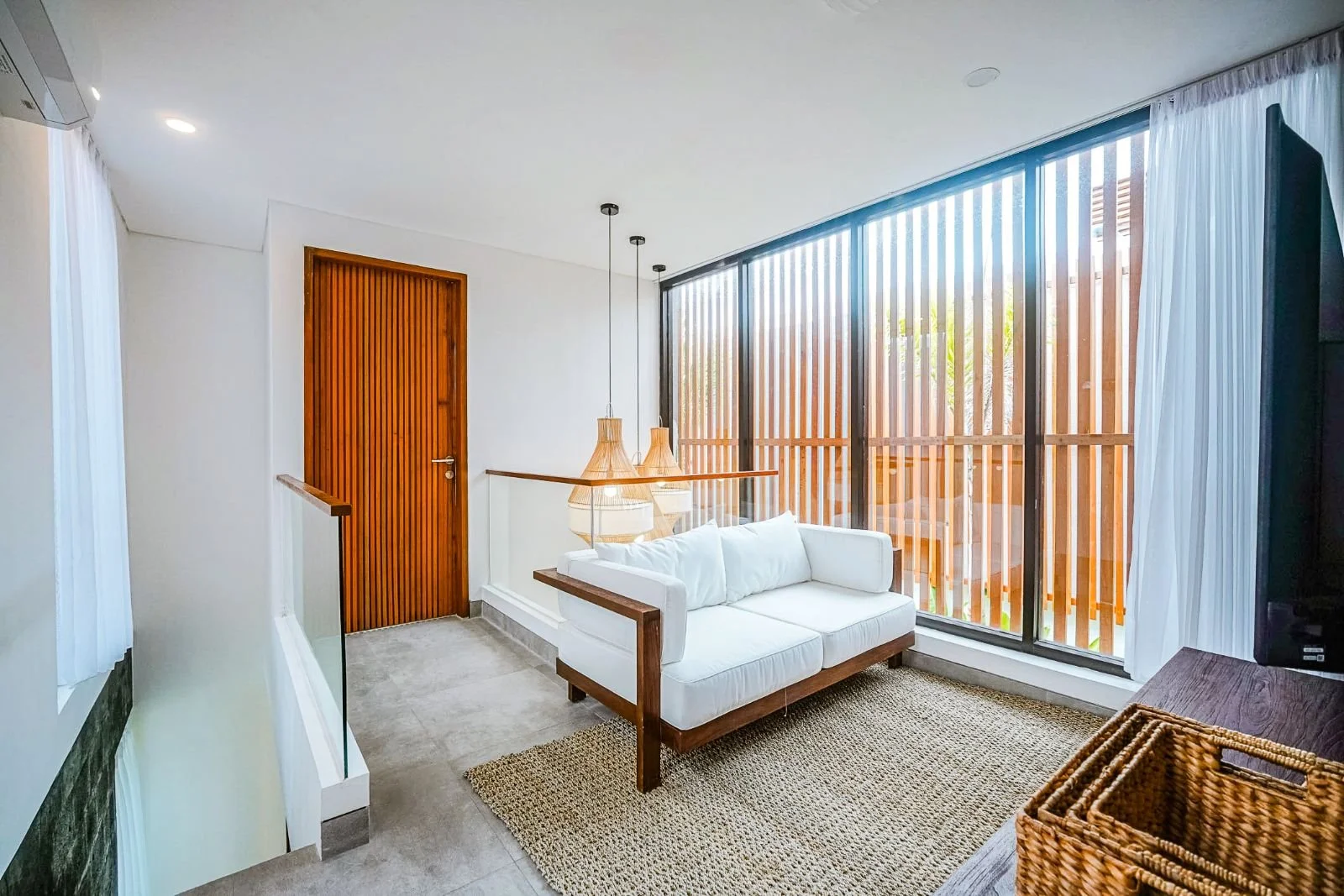 Bright living room with white sofa, wooden door, large windows with vertical wooden blinds, hanging pendant lights, beige rug, and wicker basket, overlooking an outdoor patio or garden area.