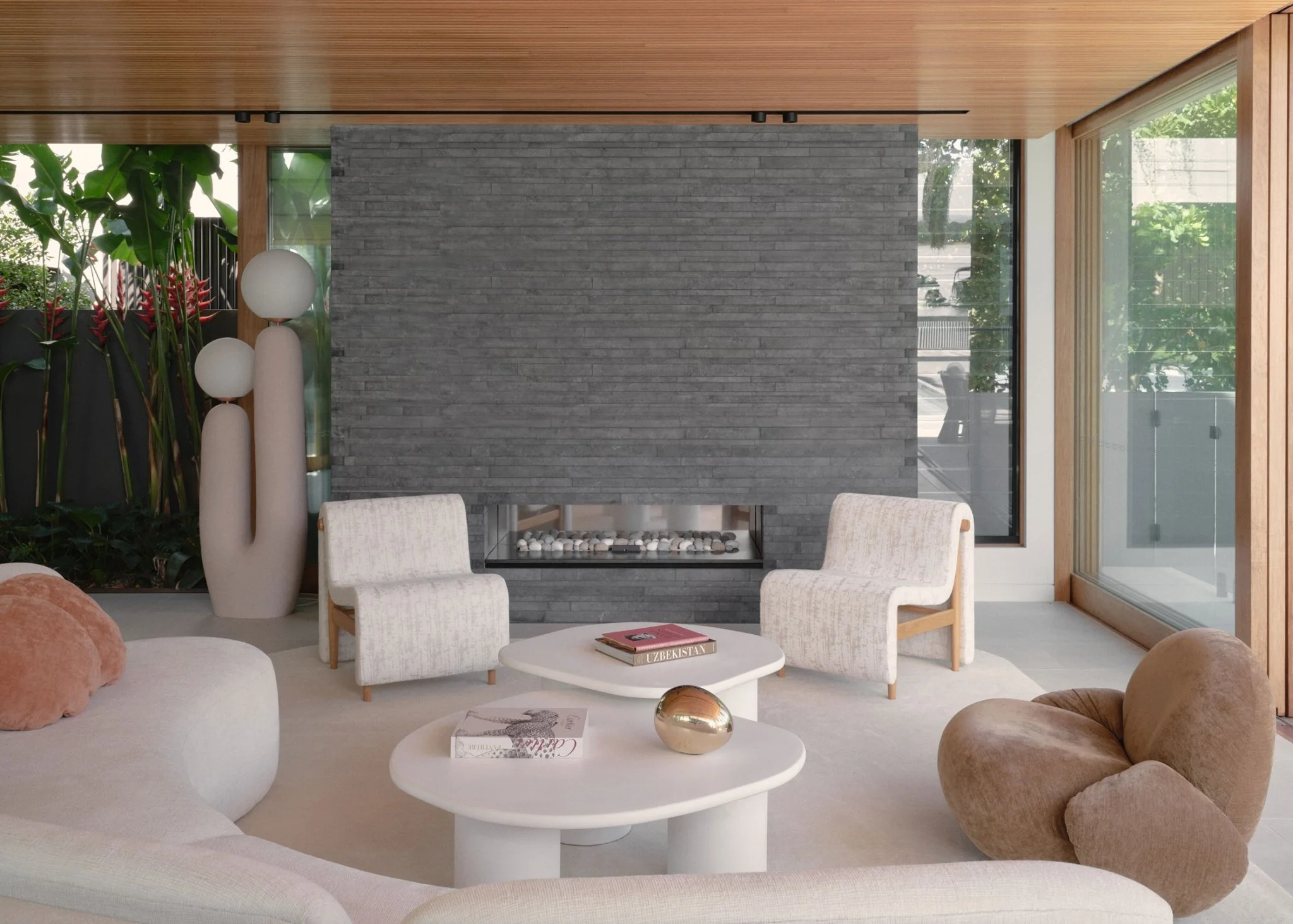 Modern living room with a gray stone fireplace, white armchairs, a white sofa, a round white coffee table with a gold decorative object and magazines, glass walls with greenery outside, and wooden ceiling and window framing.