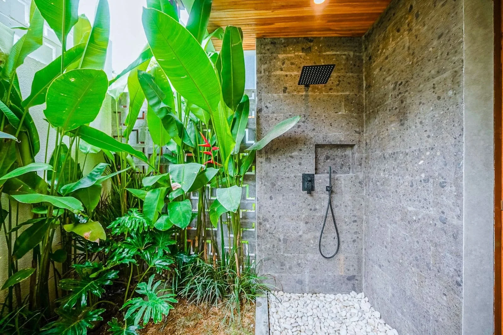 Outdoor shower with a rainfall head, hose, and a niche, surrounded by lush green tropical plants and white pebbles.