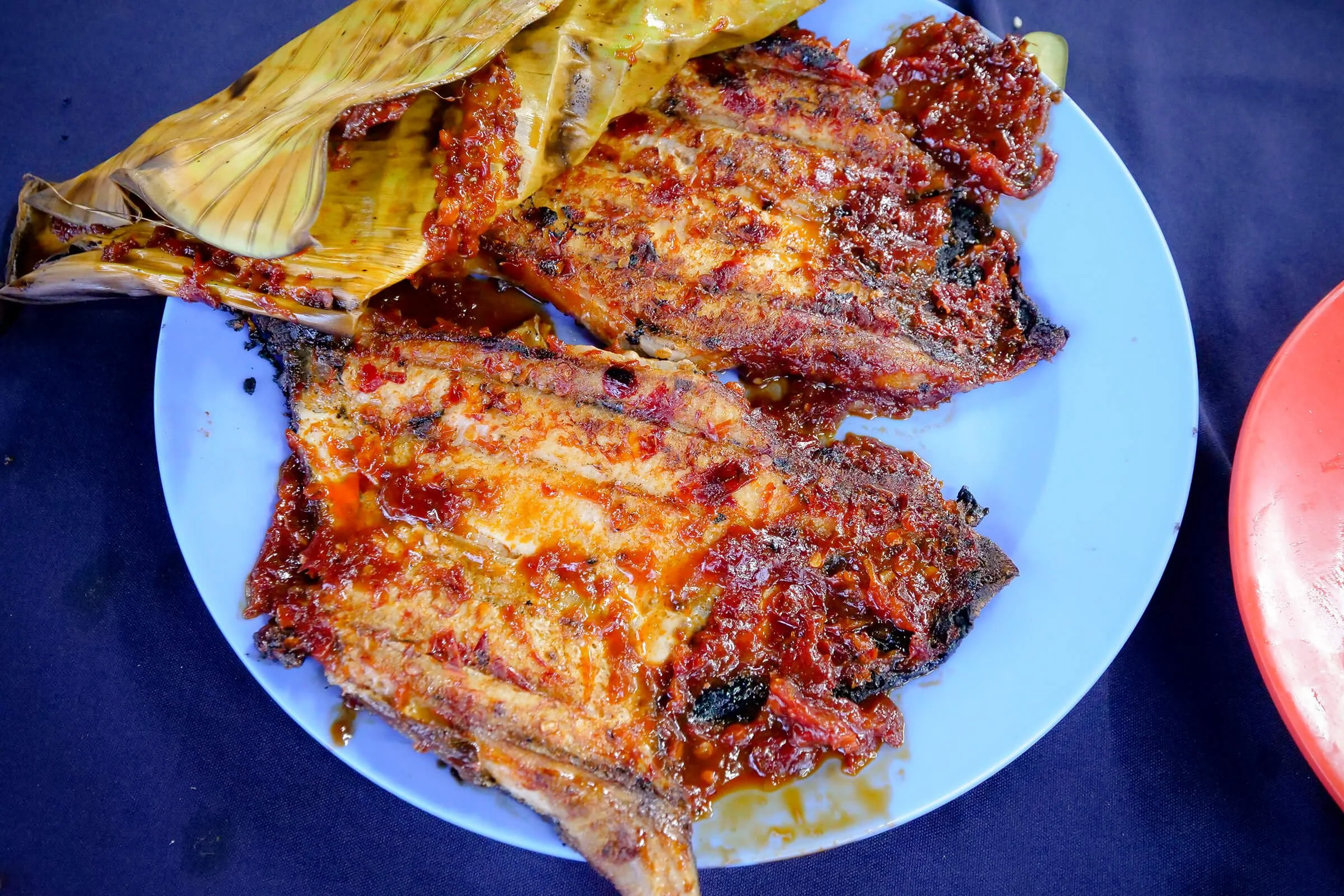 Fried fish fillets topped with tomato sauce on a white plate with a banana leaf garnish.
