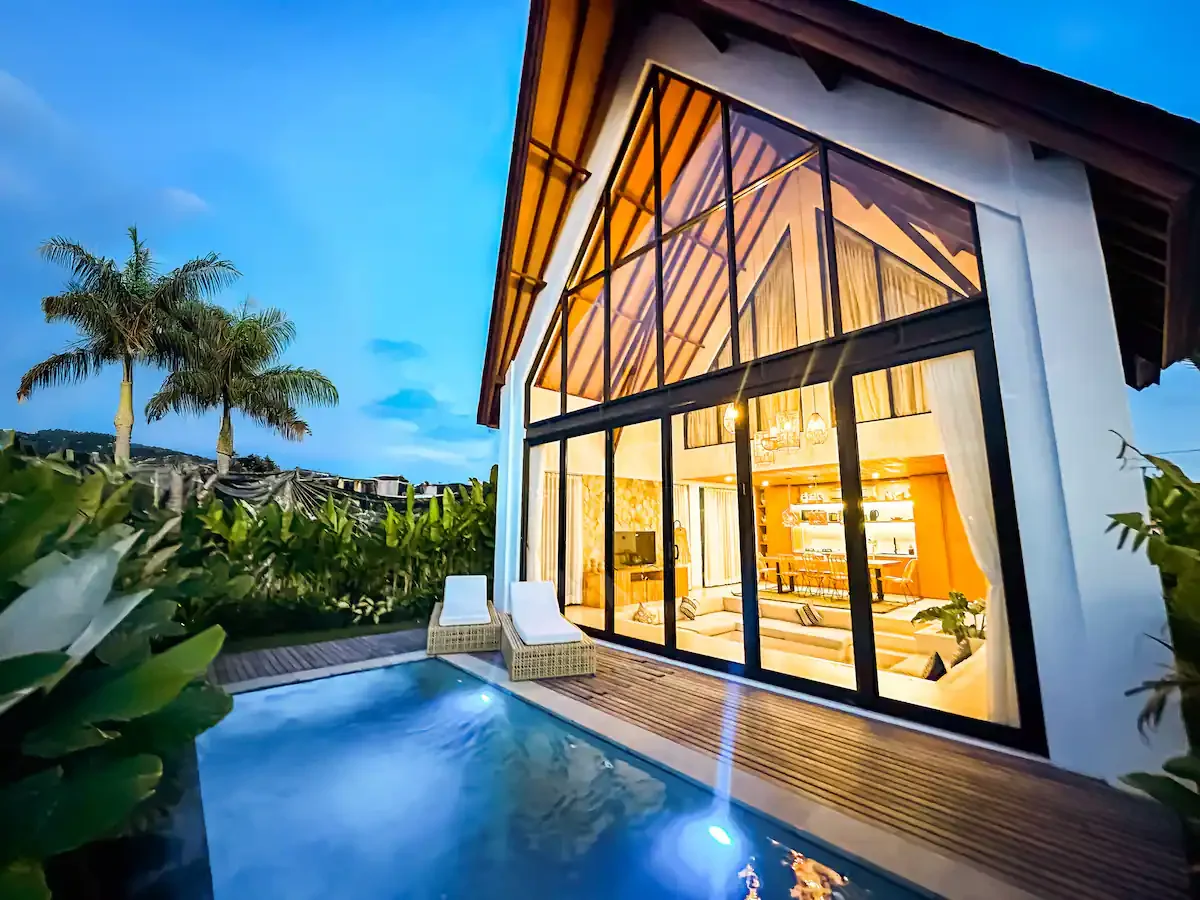 Modern house with large glass windows, outdoor pool, and palm trees at dusk, showing warm interior lighting.