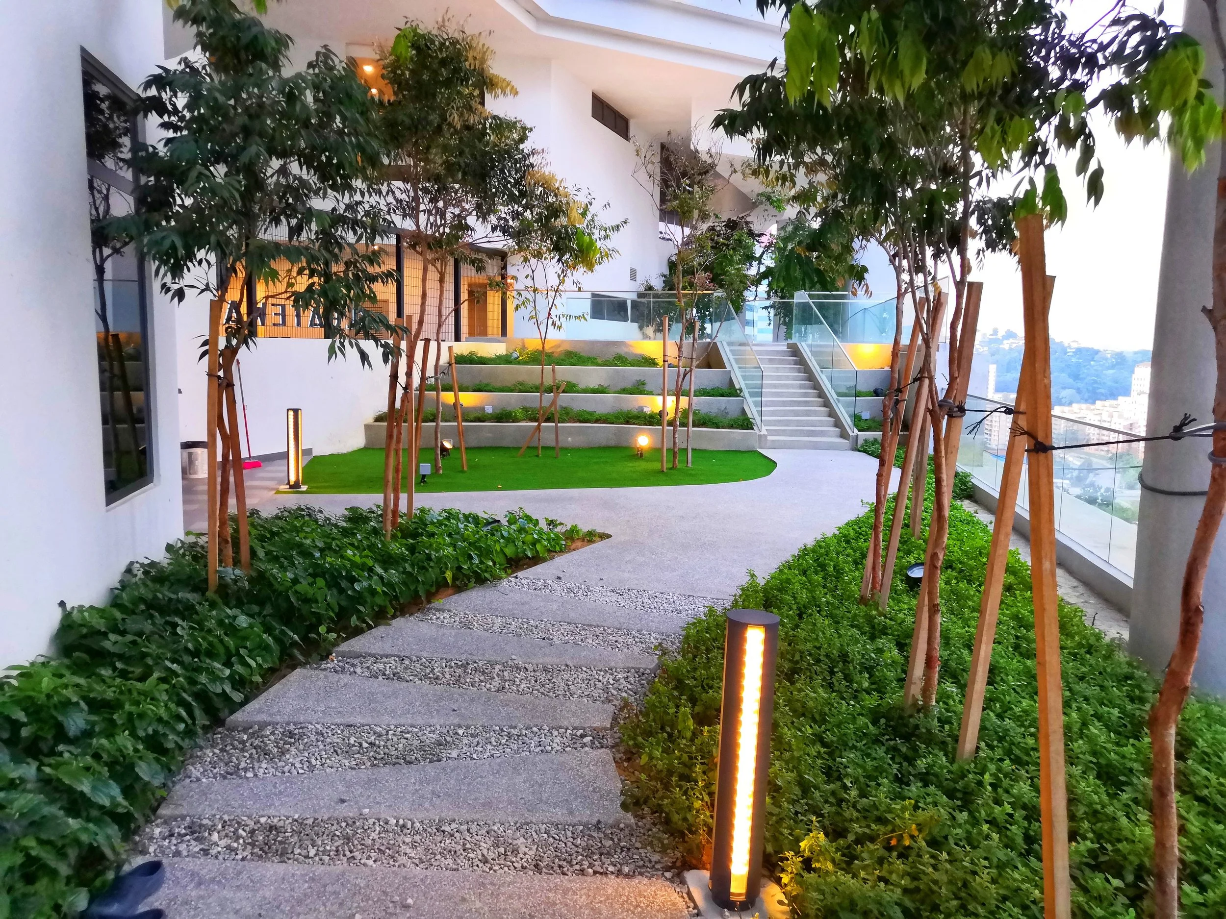 Modern rooftop garden with pathway, small trees supported by wooden stakes, green shrubs, grass, and stepping stones, illuminated by ground lights and pathway lights at dusk.